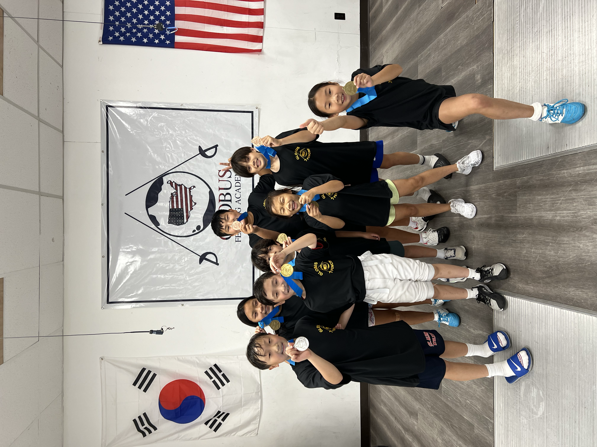 Children on a podium with medals in a martial arts competition, standing in front of flags of the United States and South Korea, with a banner for National Orthodox Academy in the background.