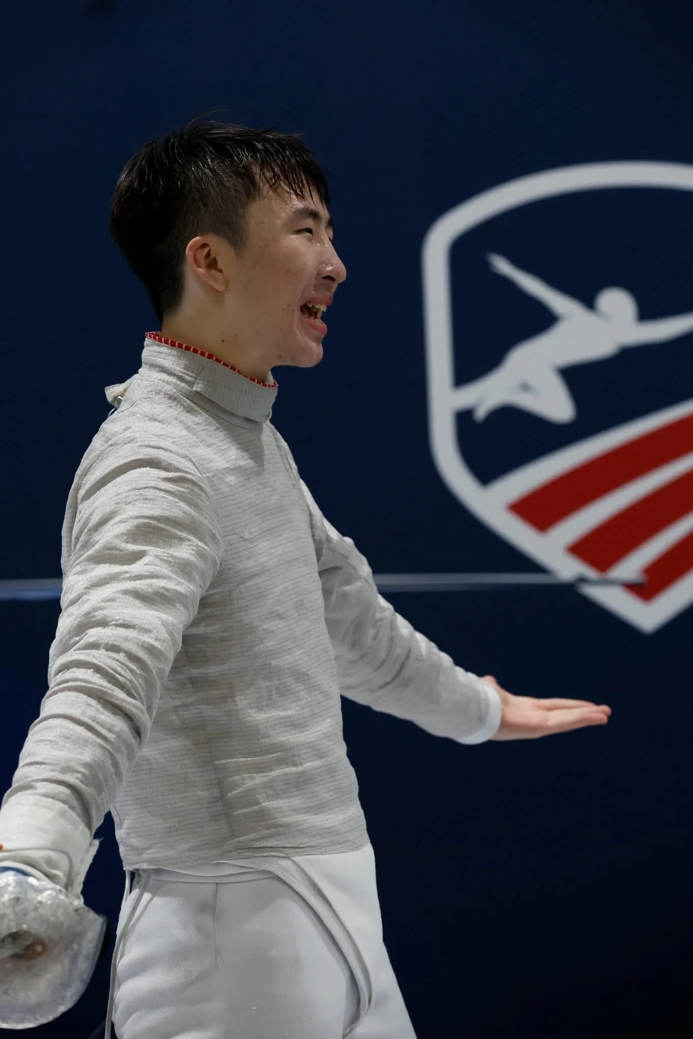 A smiling female fencer in fencing attire standing near a wall with a logo, gesturing outward with her arm.
