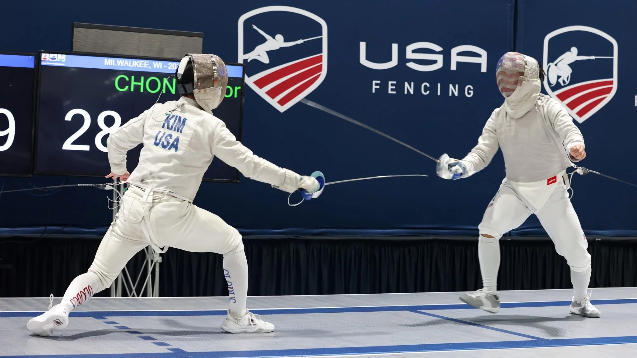 Two female fencers in white protective gear engaged in a match, swords crossed in front of a USA fencing sign.