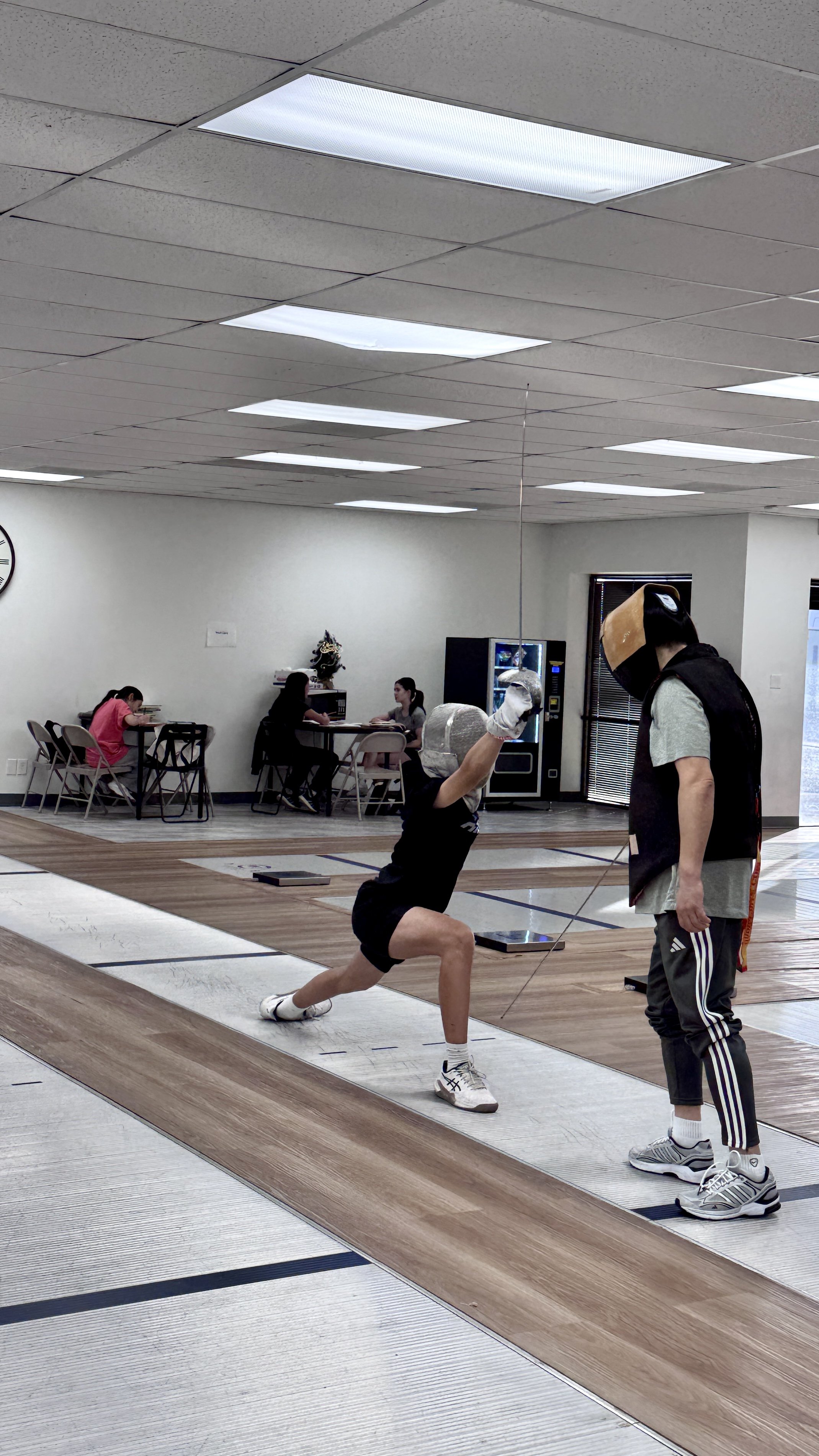 A person practicing fencing in an indoor sports facility, wearing protective gear and lunging towards another person who is holding a fencing sword and wearing a mask.