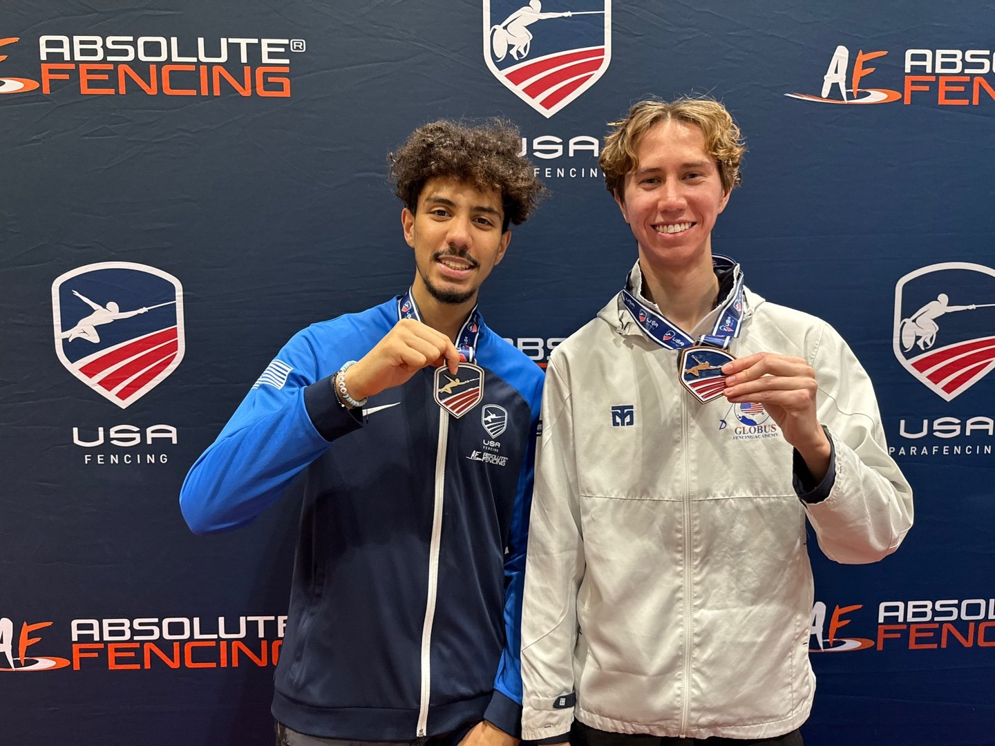 Two young men standing in front of a blue backdrop holding medals, with USA fencing logos and text in the background.