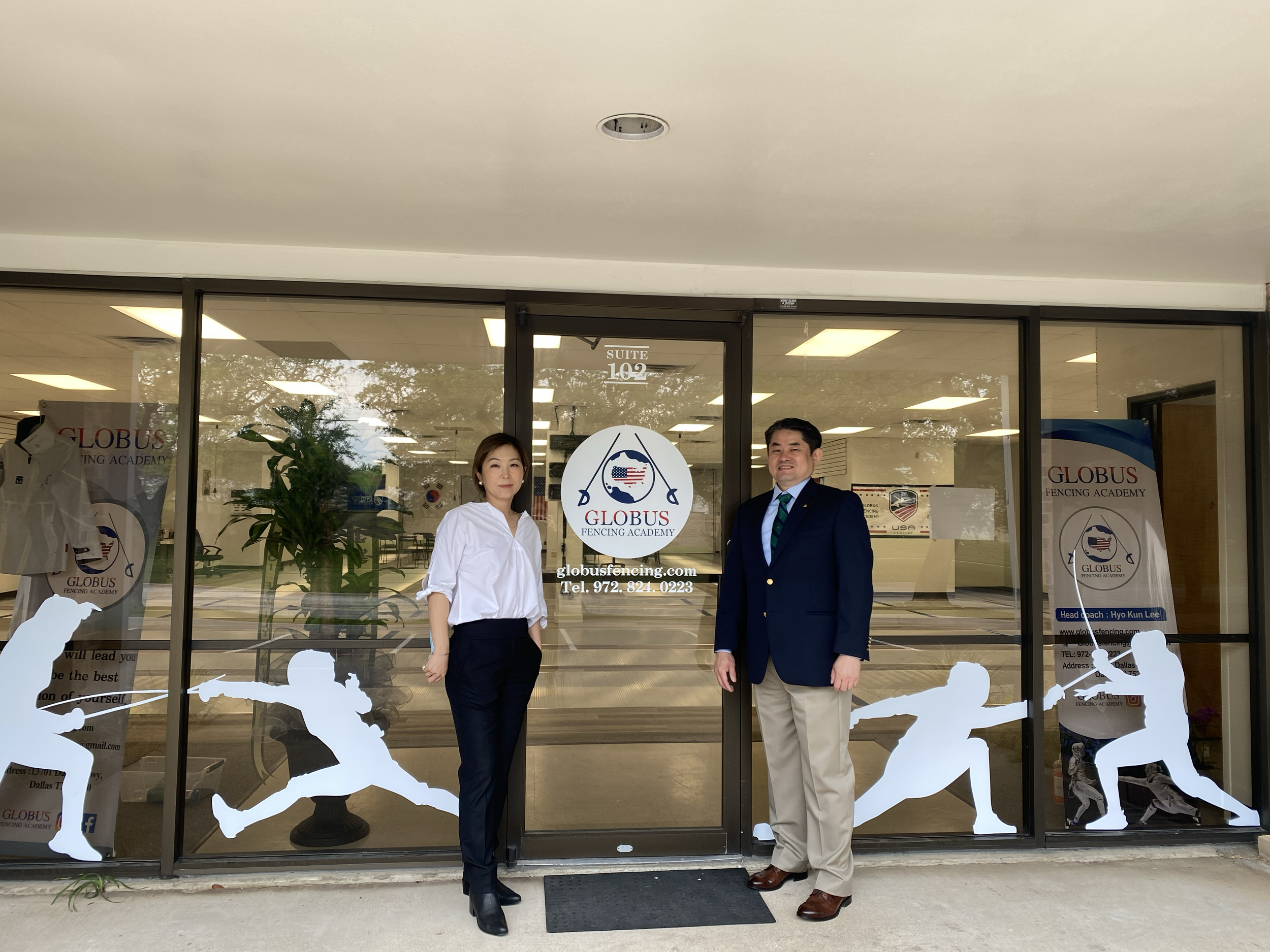 Two individuals standing outside the entrance of Globus Fencing Academy, which has glass doors with decals of fencers in various action poses. The entrance features a logo with a fencing mask and crossed foils, along with the academy's contact information. The background shows a well-lit indoor fencing area with mats and posters.