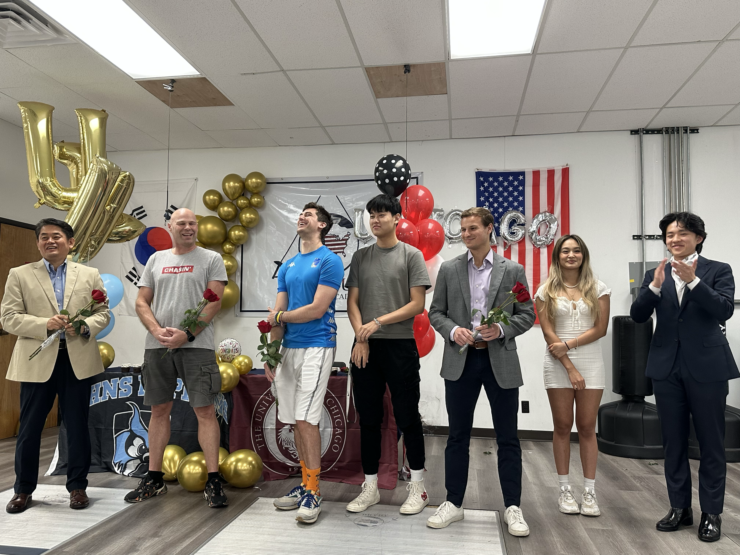 Group of people at a graduation celebration, holding roses and clapping, decorated with balloons, an American flag, and a university banner.