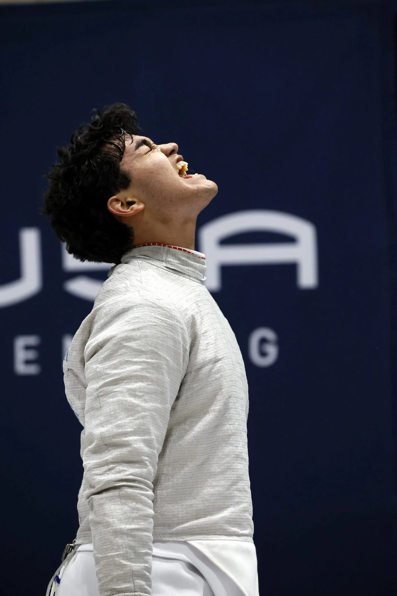 A male tennis player with short, dark, curly hair is smiling with his eyes closed and mouth open, wearing a light-colored, long-sleeved top and white shorts, on a tennis court during a match.