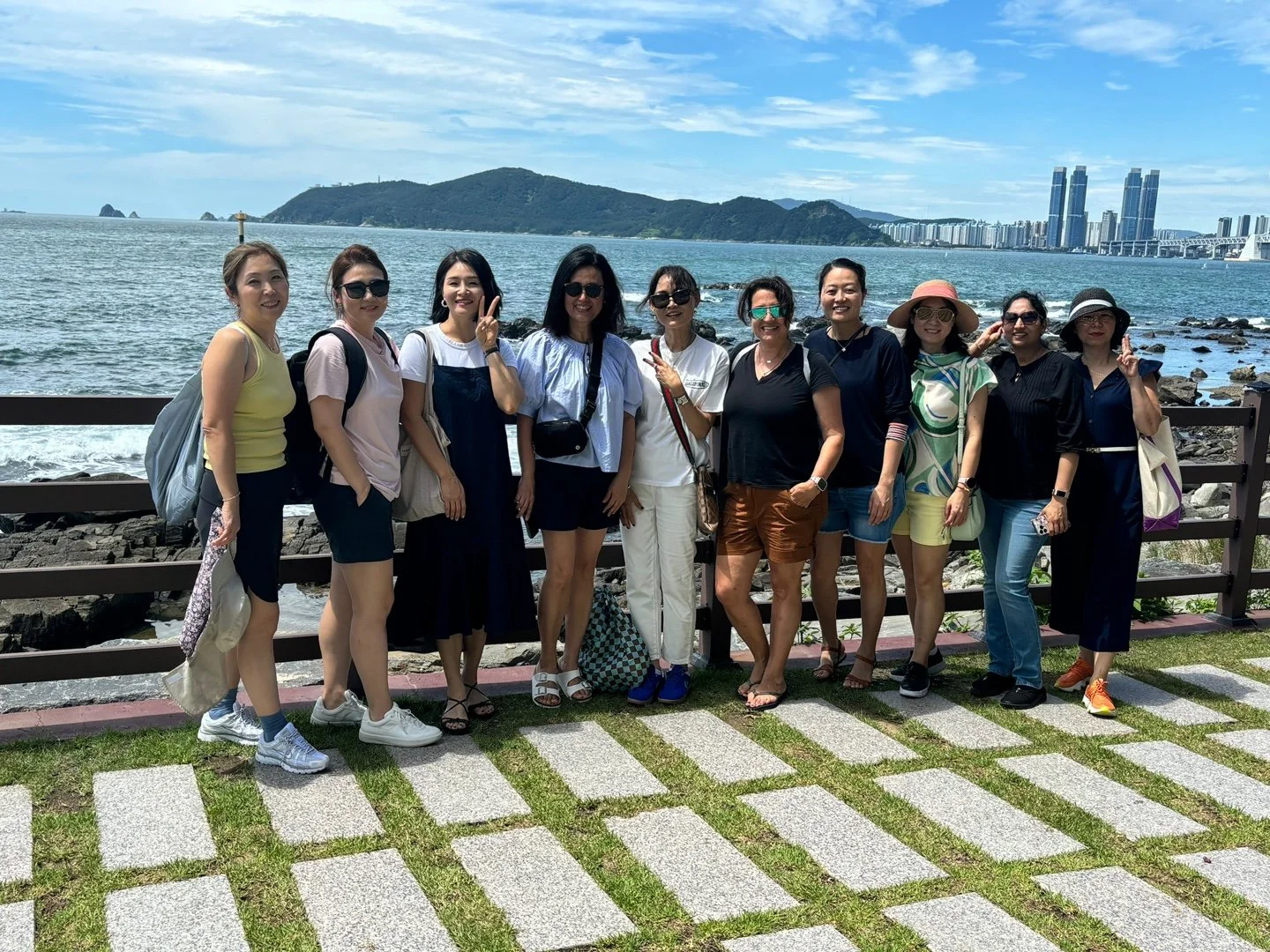 Group of eleven women standing on a waterfront walkway with a body of water, mountains, and city skyline in the background.