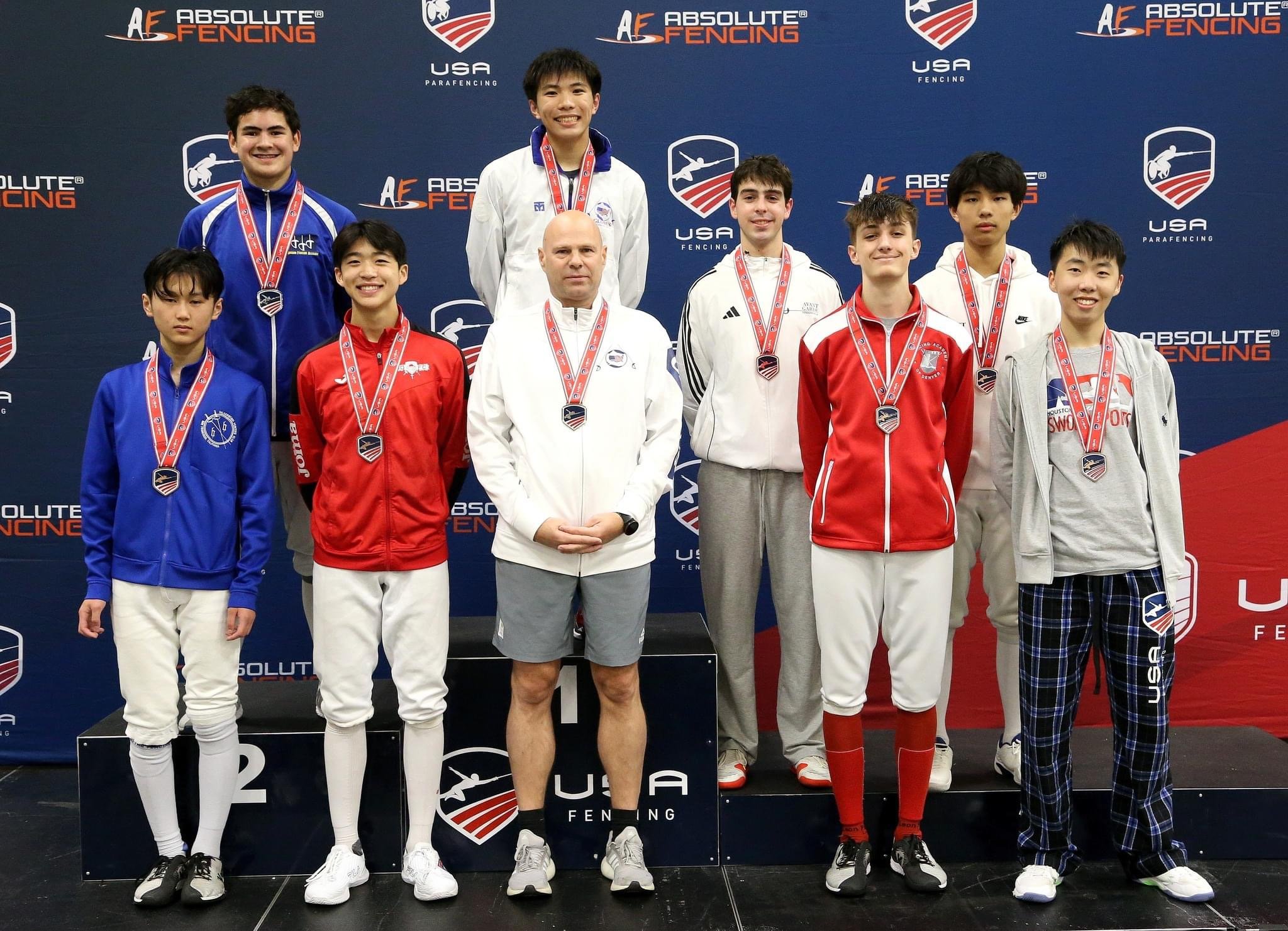 Young male fencers on the podium with medals, some wearing team jackets, in front of a backdrop with 'Absolute Fencing' and 'USA Fencing' logos.