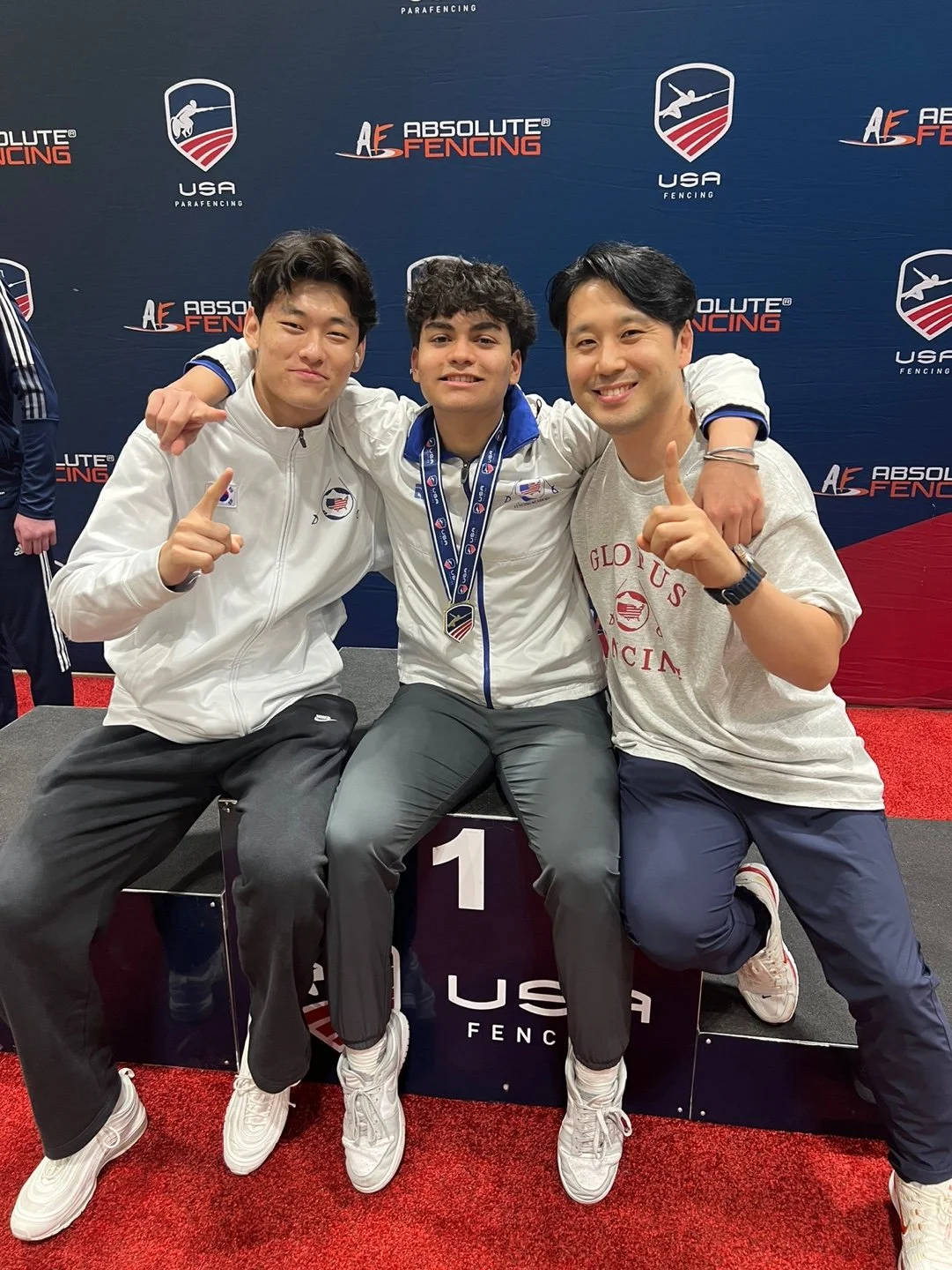 Three athletes celebrating on a winners' podium at a fencing competition, wearing medals and posing with arms around each other.