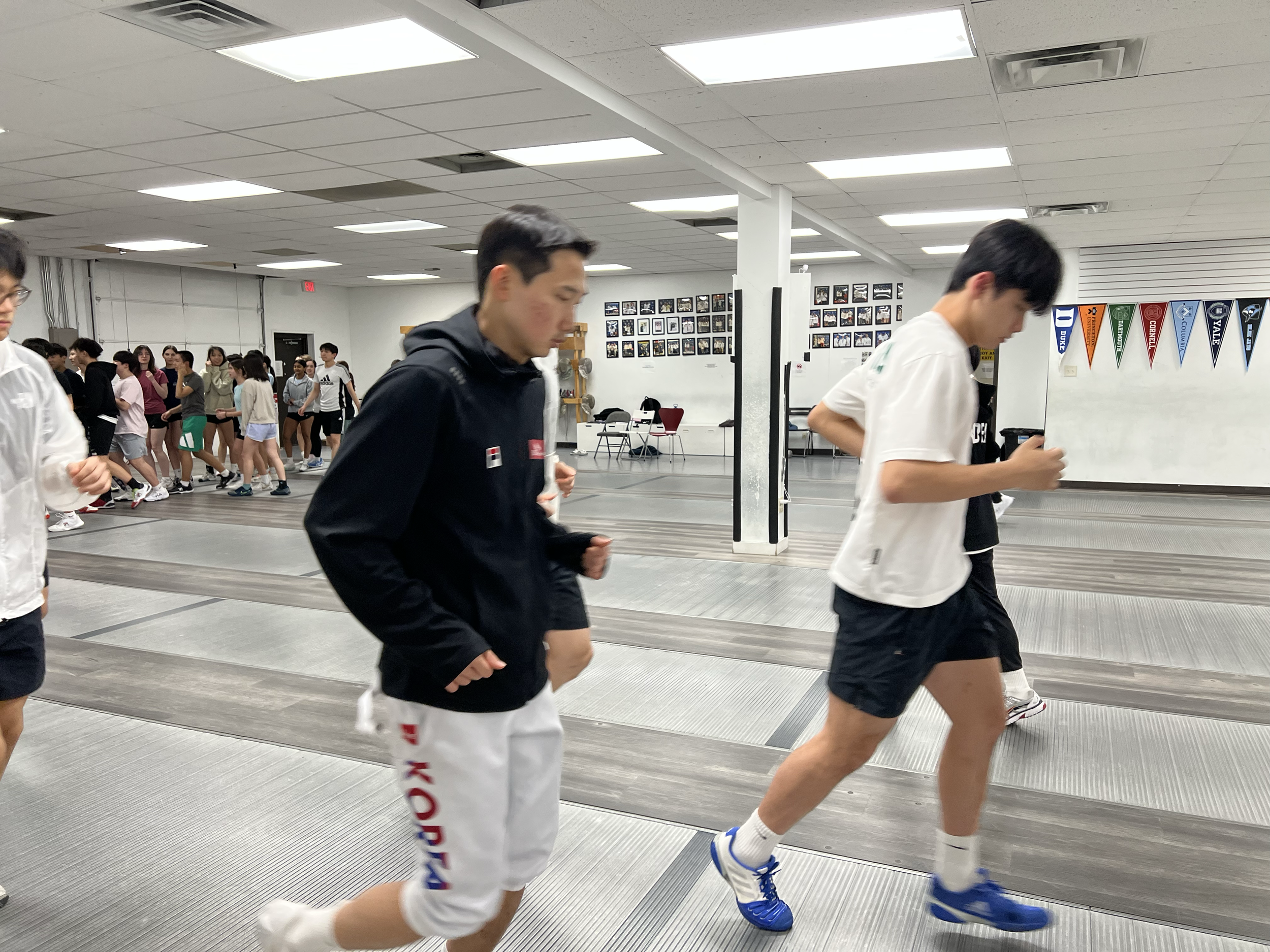 Group of people, mostly young, running on indoor fencing piste at a fencing training or event.
