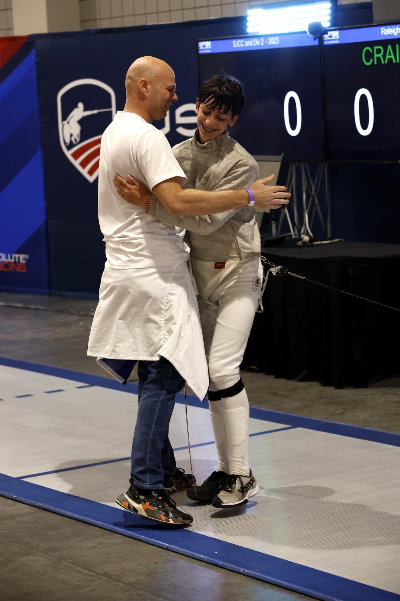 Two male fencers smiling and embracing, one in fencing gear and the other in casual clothing, on a fencing strip with a scoreboard in the background.