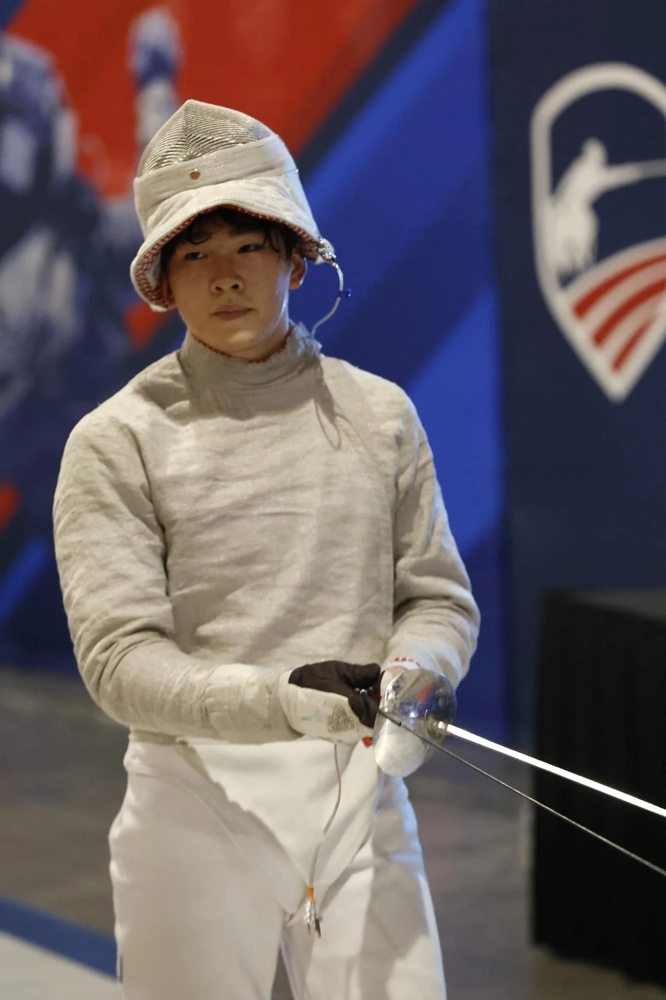 A young male fencer dressed in white fencing attire, holding a fencing sword, with fencing gear including a mask on his head, at an indoor tournament or training facility.
