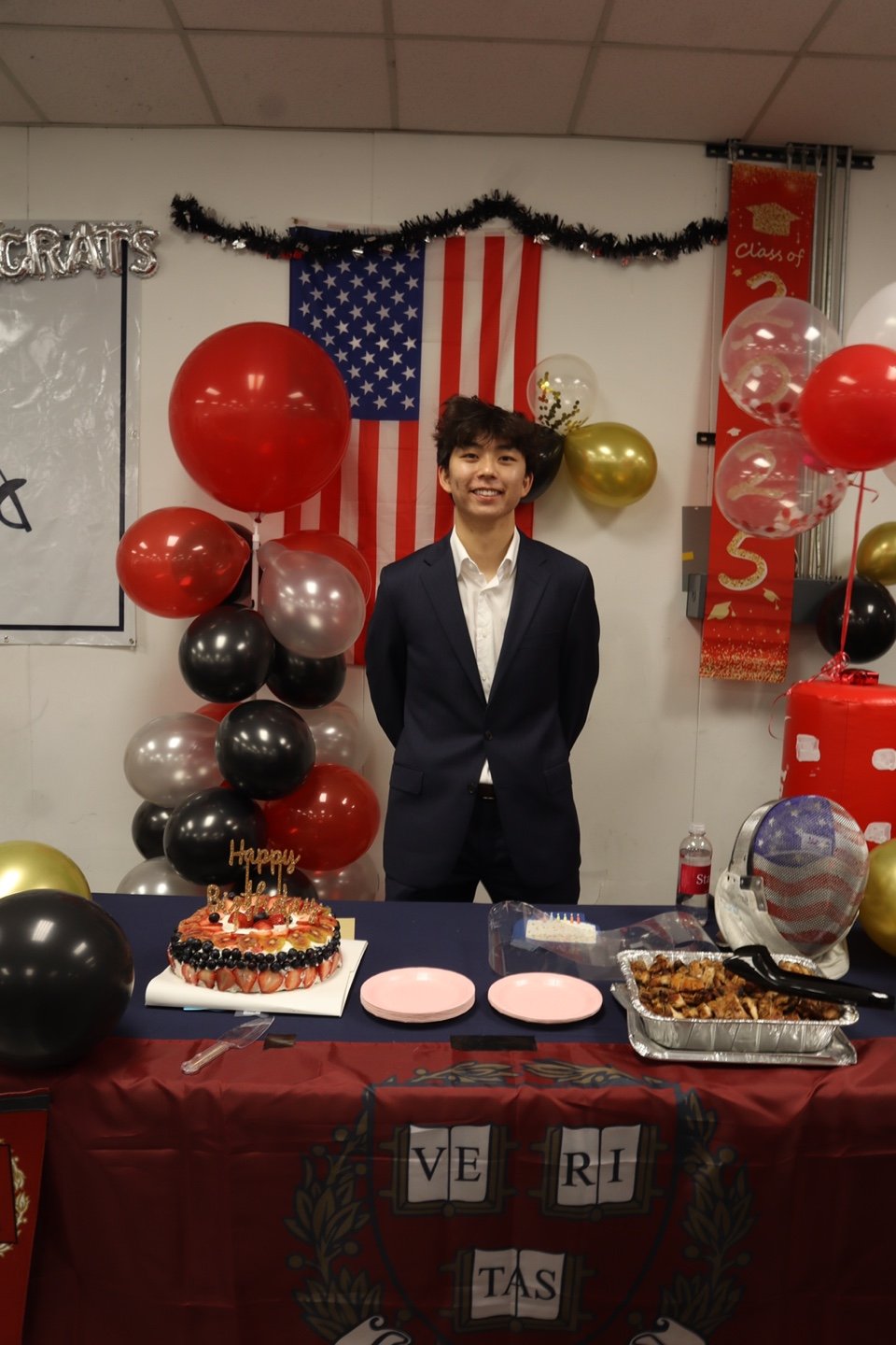 Young man in suit at a graduation celebration with American flag, balloons, birthday cake, and food table.