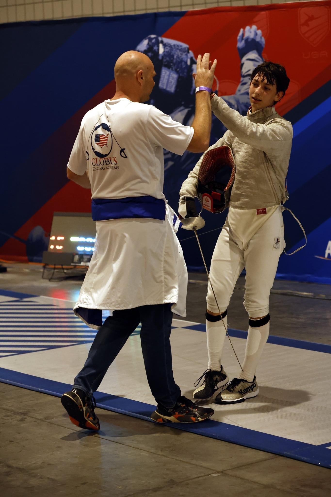 A fencing match coach giving a high five to a fencer in uniform with a fencing helmet in hand at an indoor sports arena.