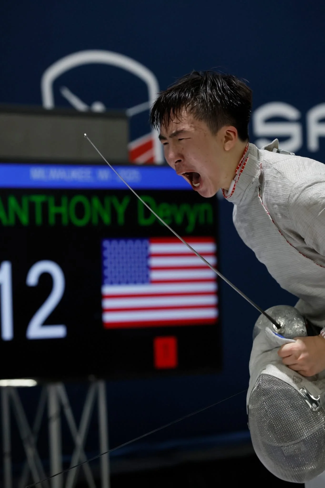 A male fencer in a white uniform reacts passionately during a fencing match, holding his mask in his left hand. The scoreboard in the background displays his name and an American flag.