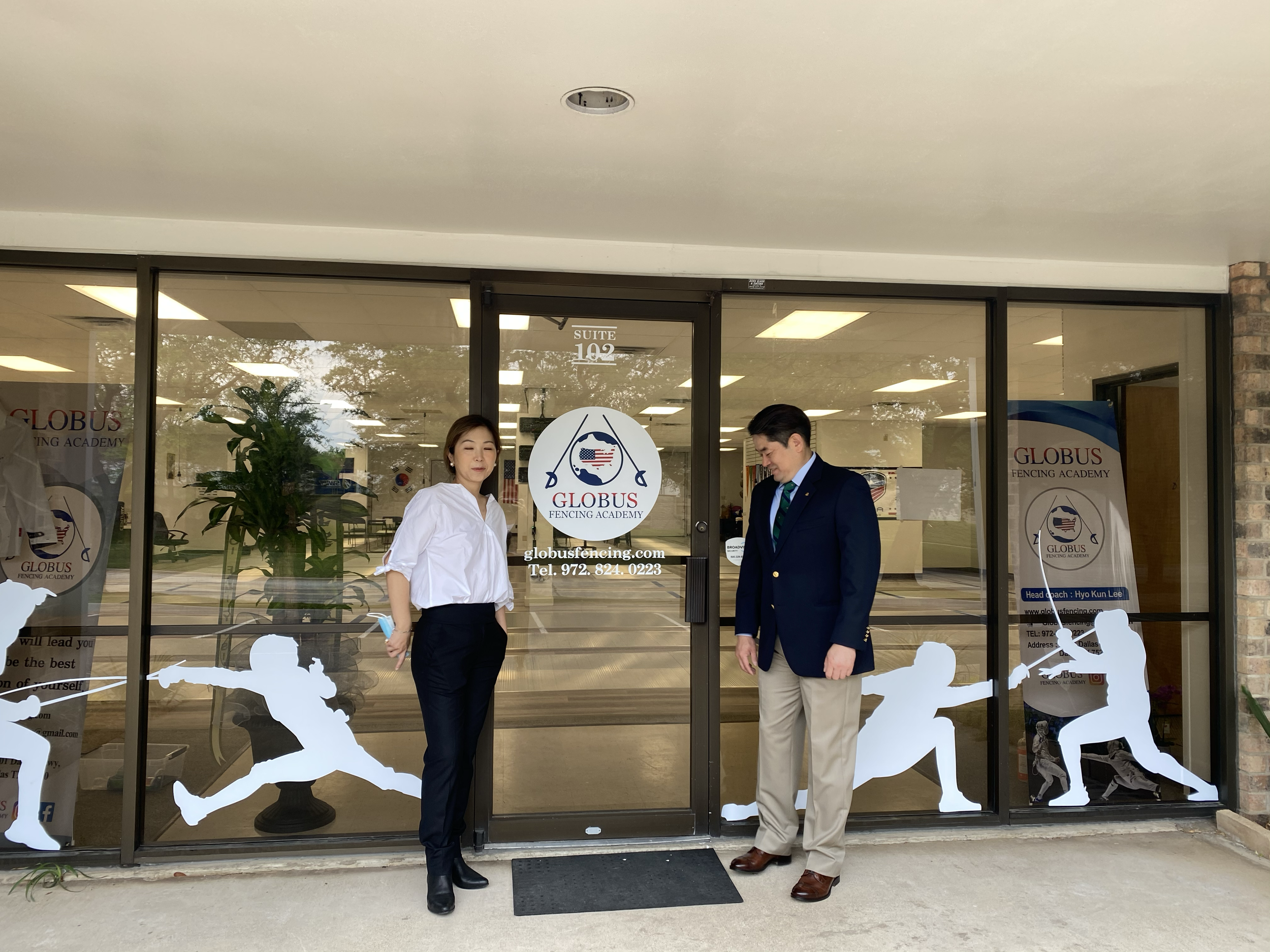 A man and a woman standing outside a fencing academy, in front of a glass door with the academy's logo. The logo features a Union Jack, an American flag, and fencing swords. The woman is dressed in a white blouse and dark pants, holding a face mask i