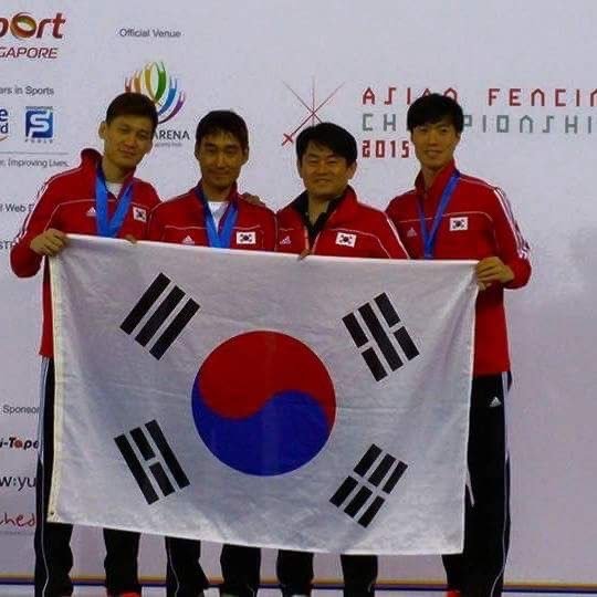 Four athletes in red uniforms standing on a podium holding the South Korean flag with a white background and event banners, celebrating at a sports championship.
