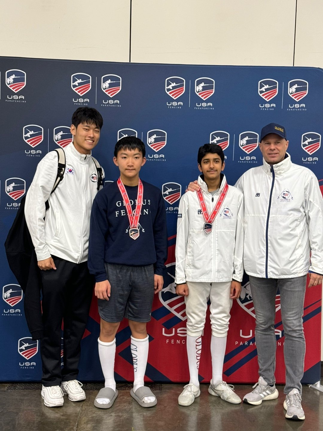 Four individuals, two teenagers and two adults, standing in front of a USA fencing backdrop. The two teenagers in the middle are wearing medals around their necks, and all are dressed in athletic and fencing-related attire.