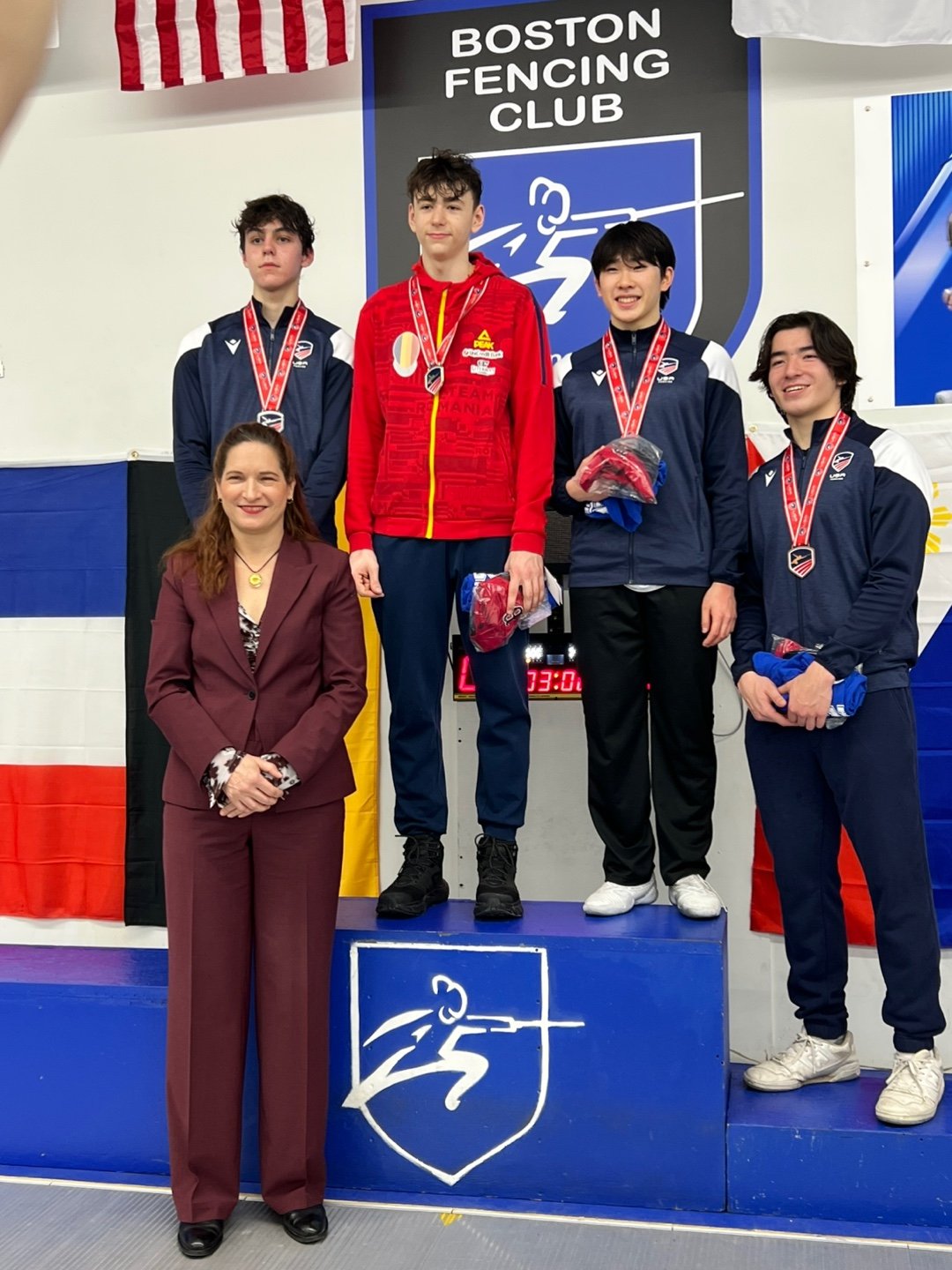 A group of young athletes standing on a winners' podium at a fencing event, with medals around their necks, and a woman in a burgundy suit standing in front of the podium. The background shows a sign for the Boston Fencing Club and flags.