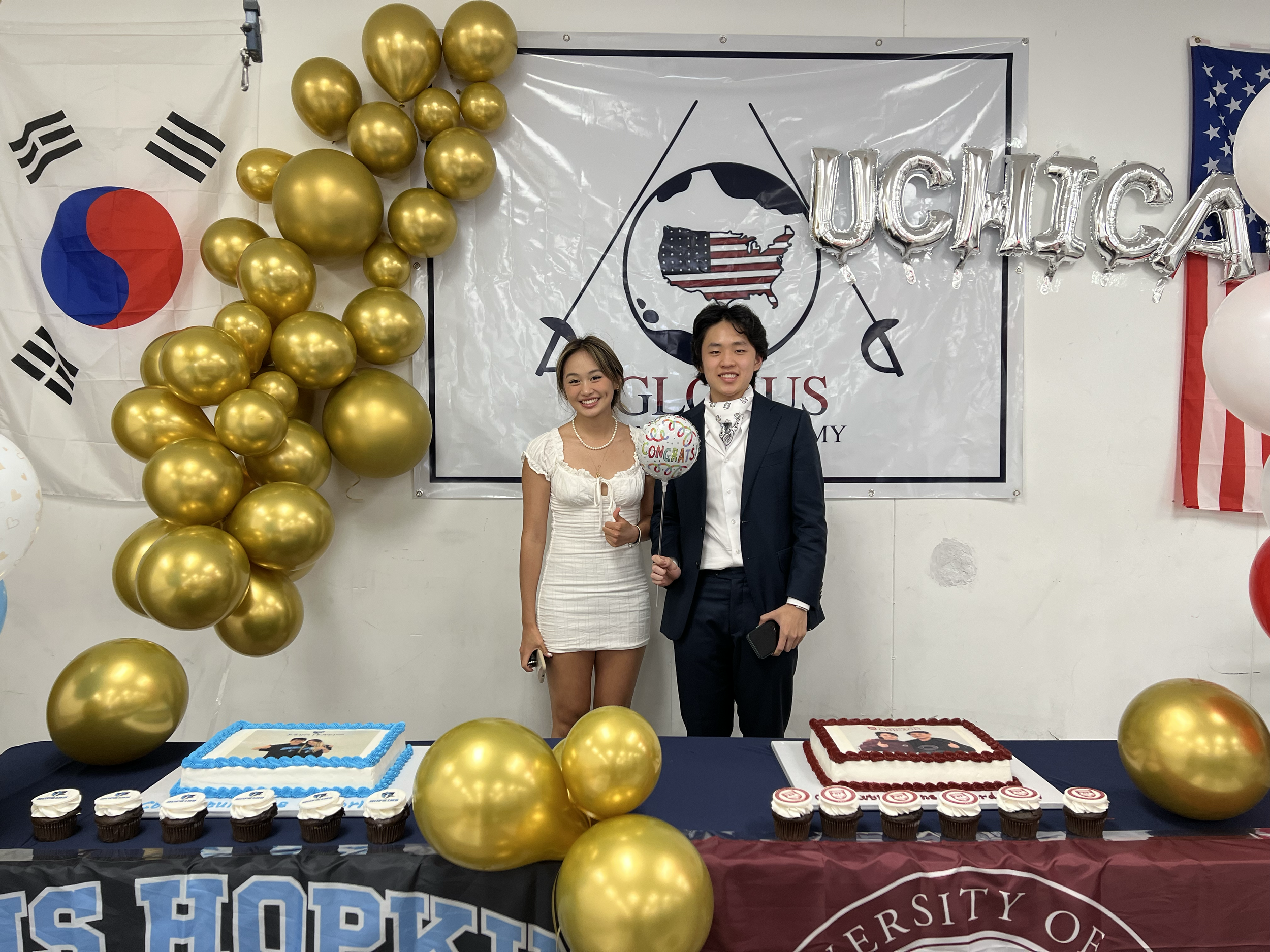 Two young people stand behind a table with cakes and cupcakes, celebrating a graduation. The woman wears a white dress, and the man wears a dark suit. Decorated balloons, including gold ones, and flags of South Korea and the USA are in the background