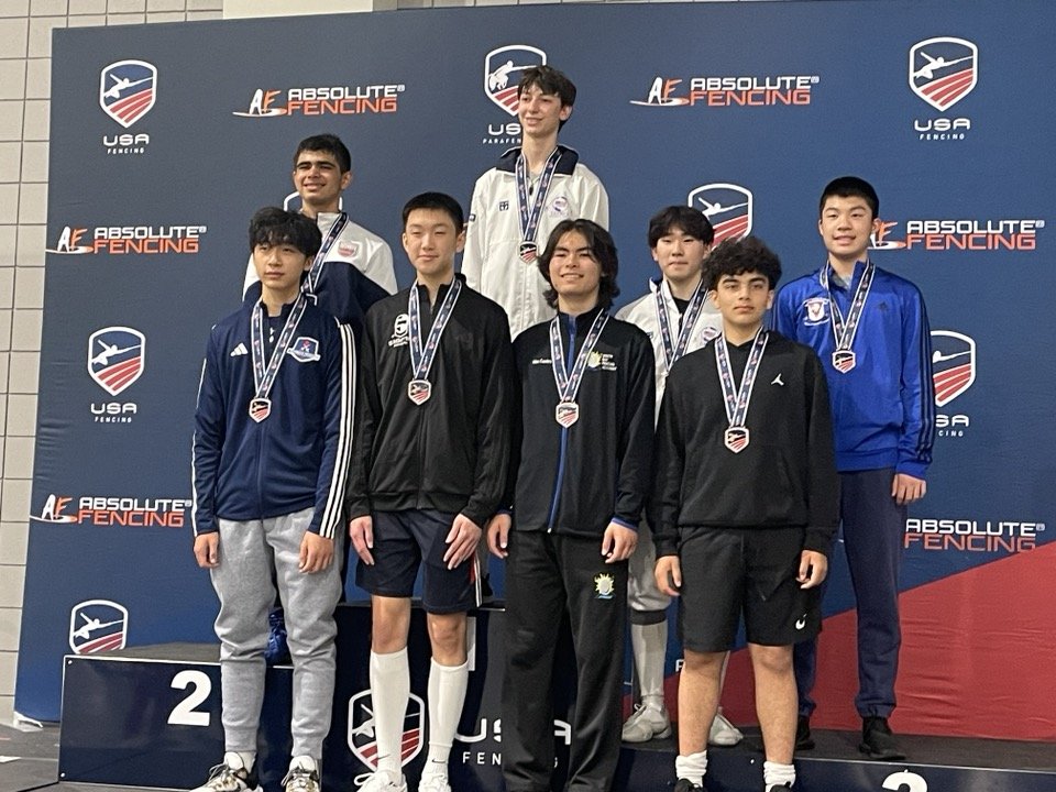 Group of young athletes standing on a podium with medals, at a fencing competition awards ceremony, with a backdrop displaying 'Absolute Fencing' and 'USA Fencing' logos.