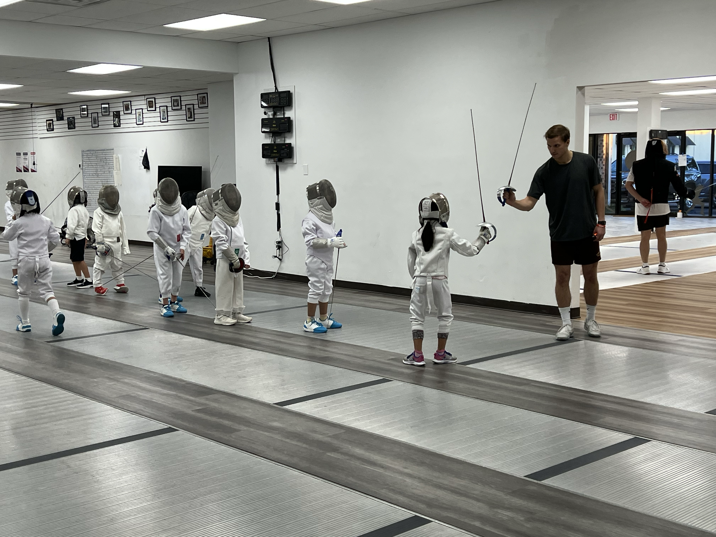 Children in fencing gear practicing fencing with their instructor in a gymnasium.