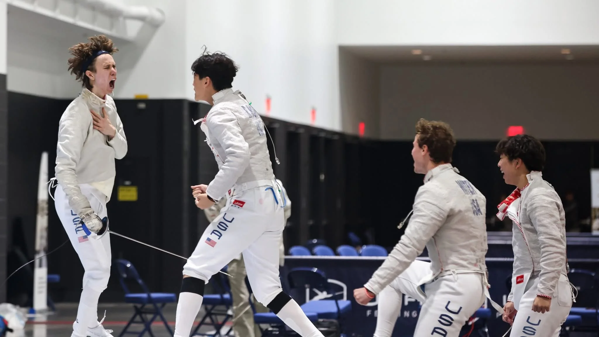 A group of five Olympic fencers in white uniforms celebrating on the fencing strip, with one woman visibly emotional, clutching her chest, and four men smiling and congratulating each other.