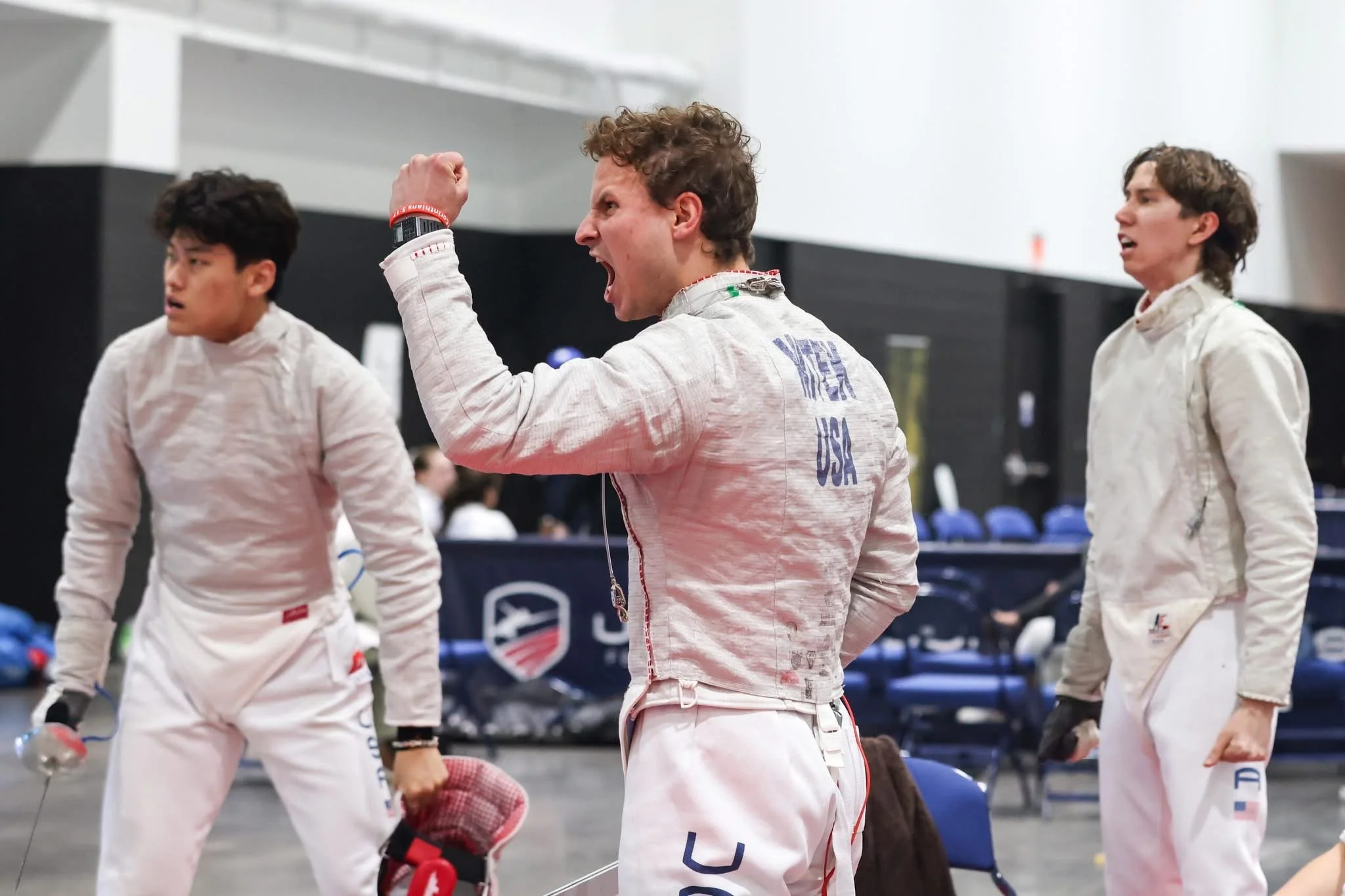 Three male fencers in fencing gear in an indoor fencing area, one celebrating with clenched fist, others looking surprised or focused.