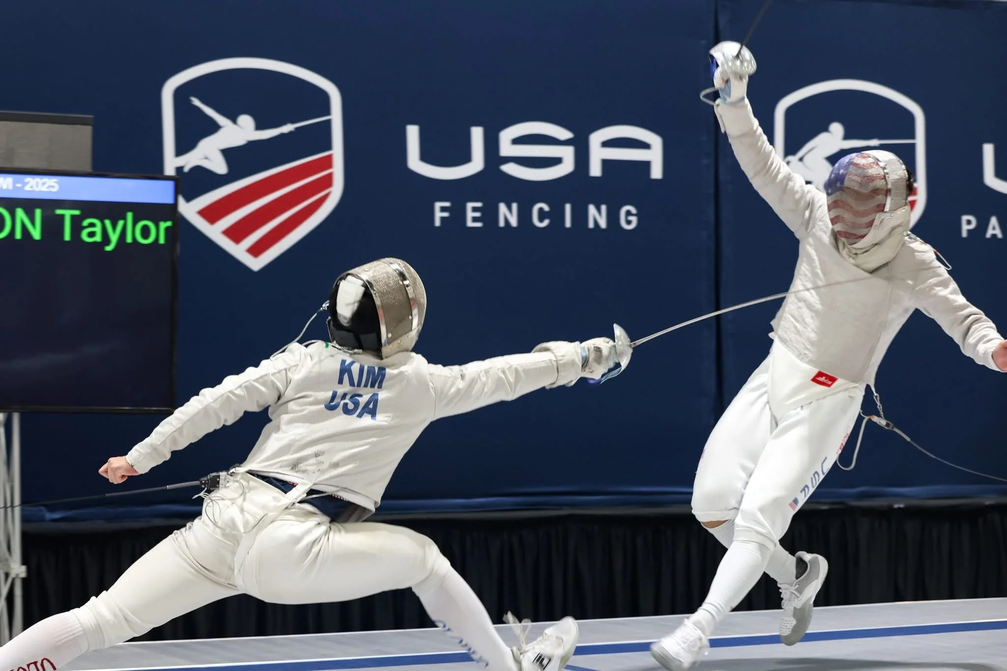 Two fencers in white protective gear practicing at an indoor fencing competition, with one lunging and the other recoiling, against a blue backdrop with USA Fencing logos.