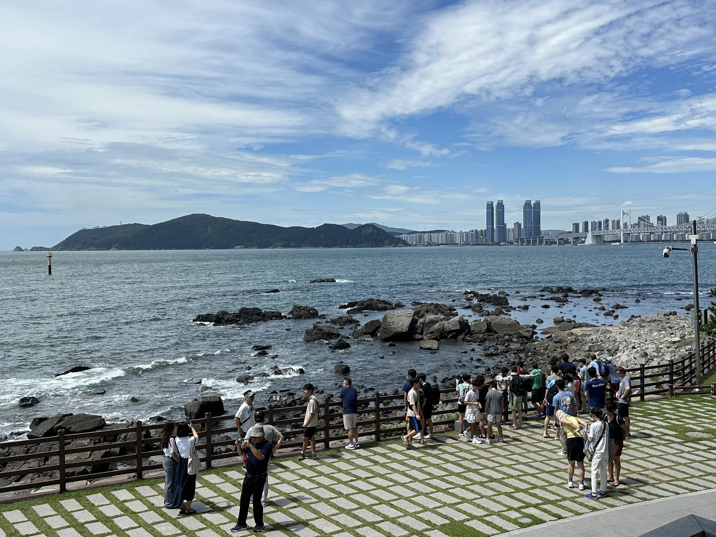 People walking and taking photos along a rocky coastline with a city skyline, with tall modern buildings, in the background on a sunny day.