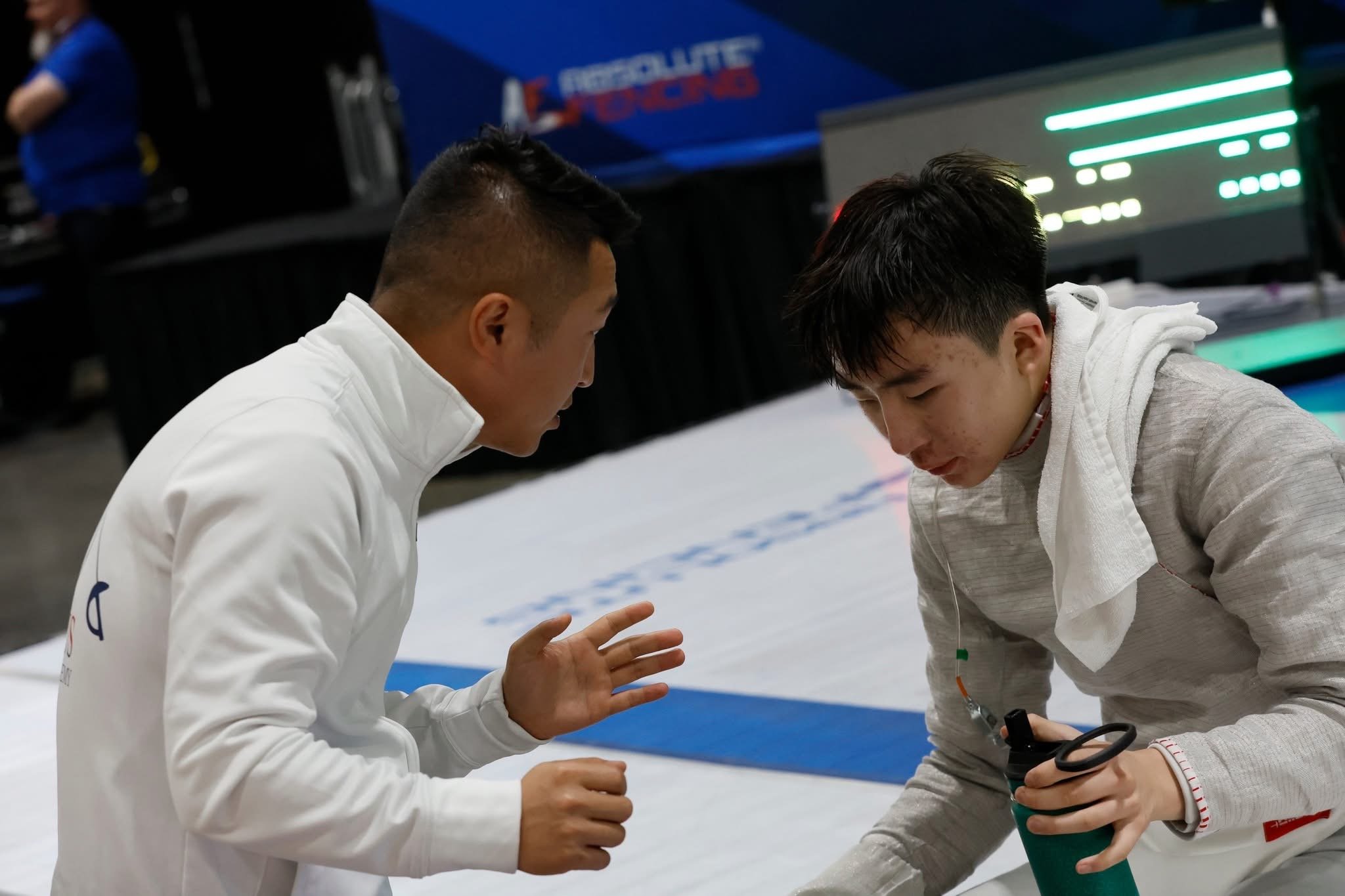 Two young male athletes in white sports jackets engage in a discussion on a sports field, with one holding a green water bottle, during an indoor sports event.