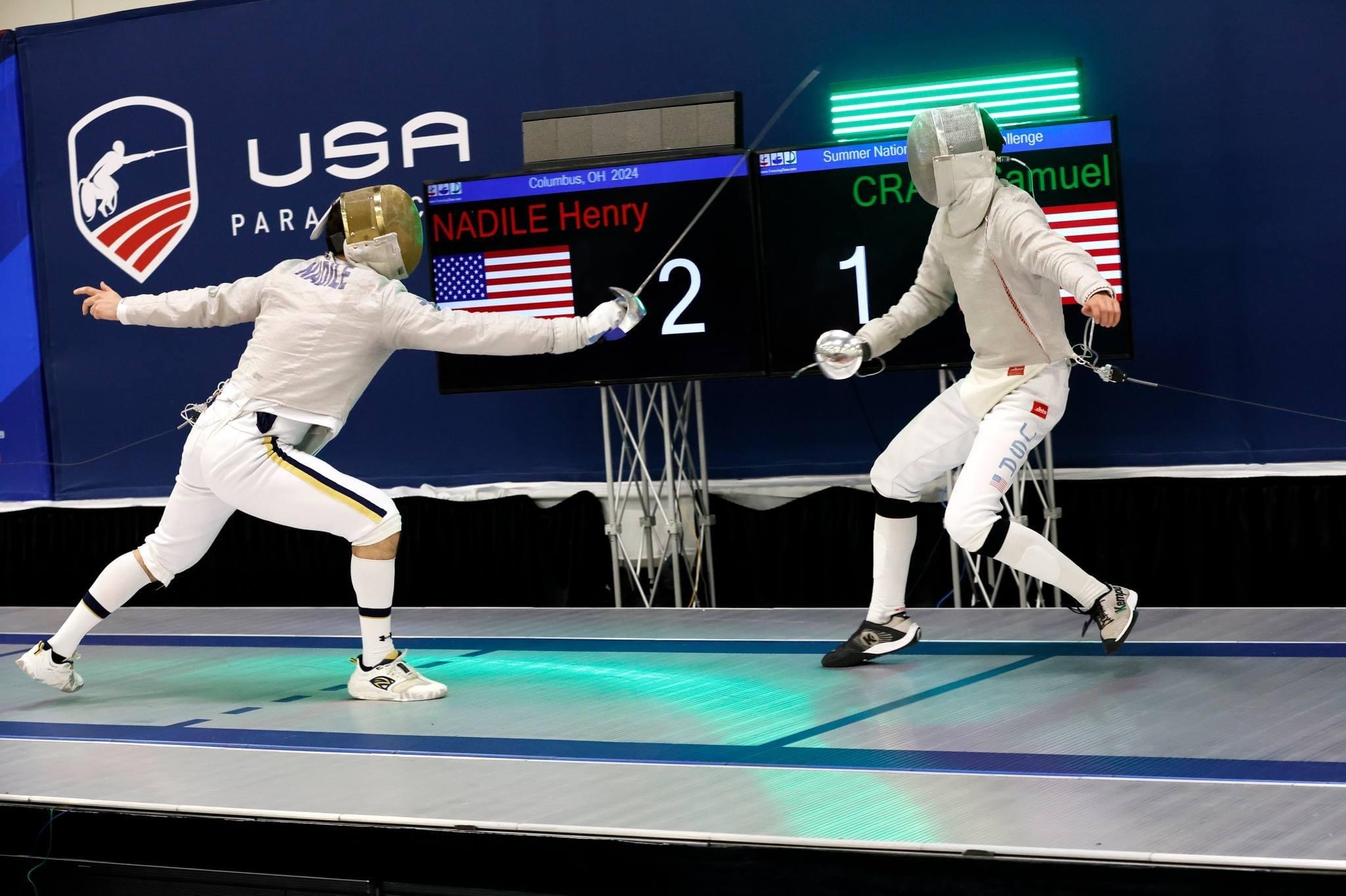 Two American male foil fencers in full gear duel on a metallic strip during a competition, with a scoreboard displaying their names and scores in the background.