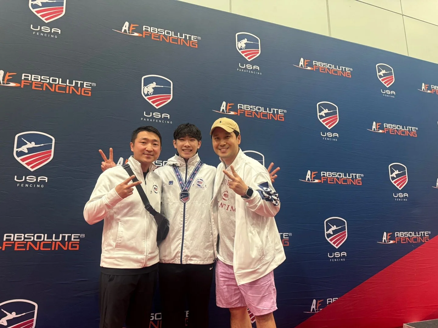 Three men standing in front of a backdrop featuring logos for USA Fencing and Absolute Fencing. The man in the middle is smiling and wearing a medal around his neck. The men on either side are making peace signs with their hands.