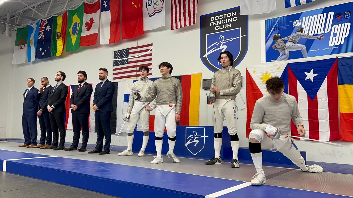 Fencers dressed in white gear standing and kneeling on a competition podium, with national flags and banners of the Boston Fencing Club and World Cup in the background.