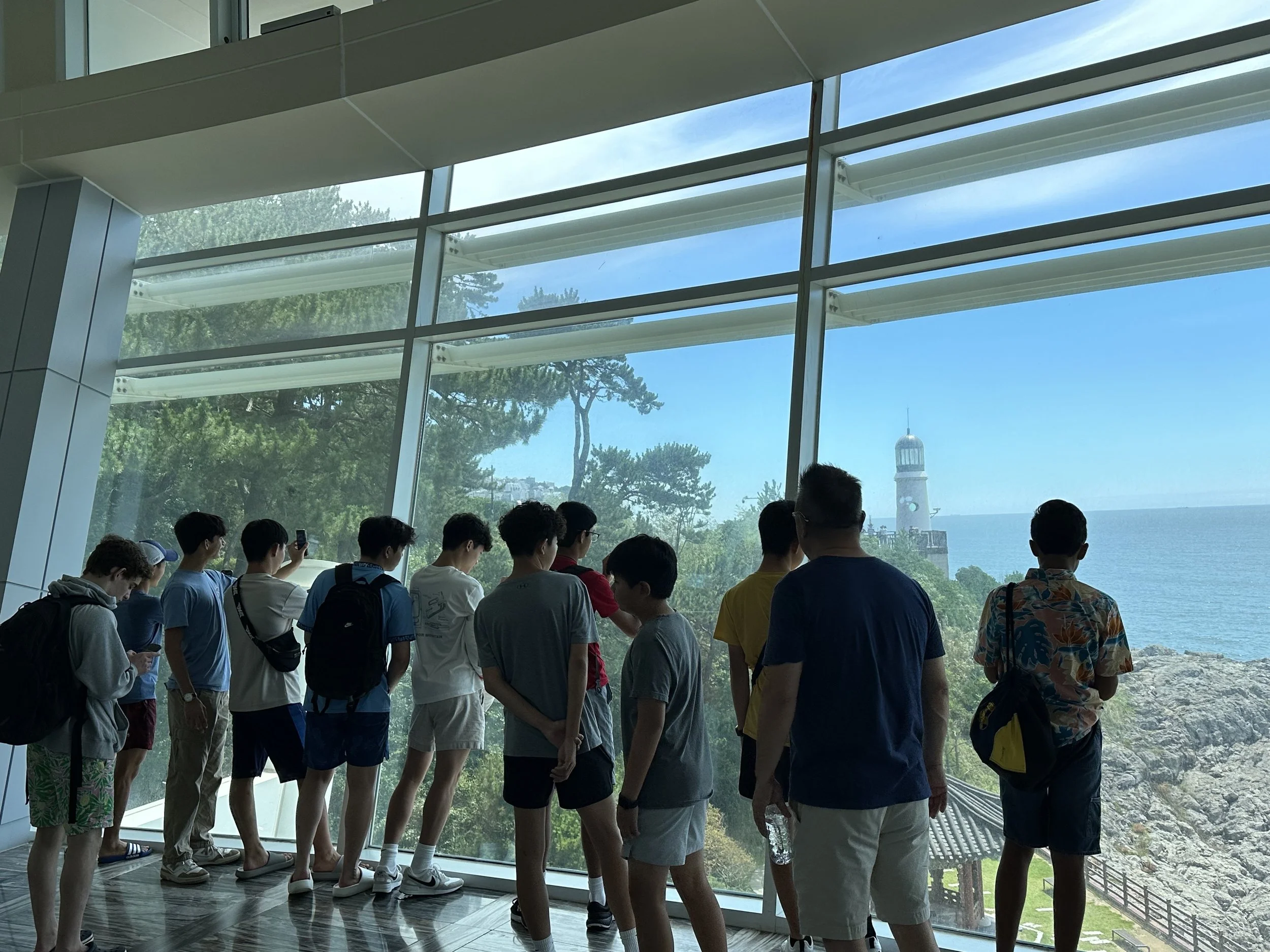Group of young people looking out a large glass window at a lighthouse on a rocky coastline.