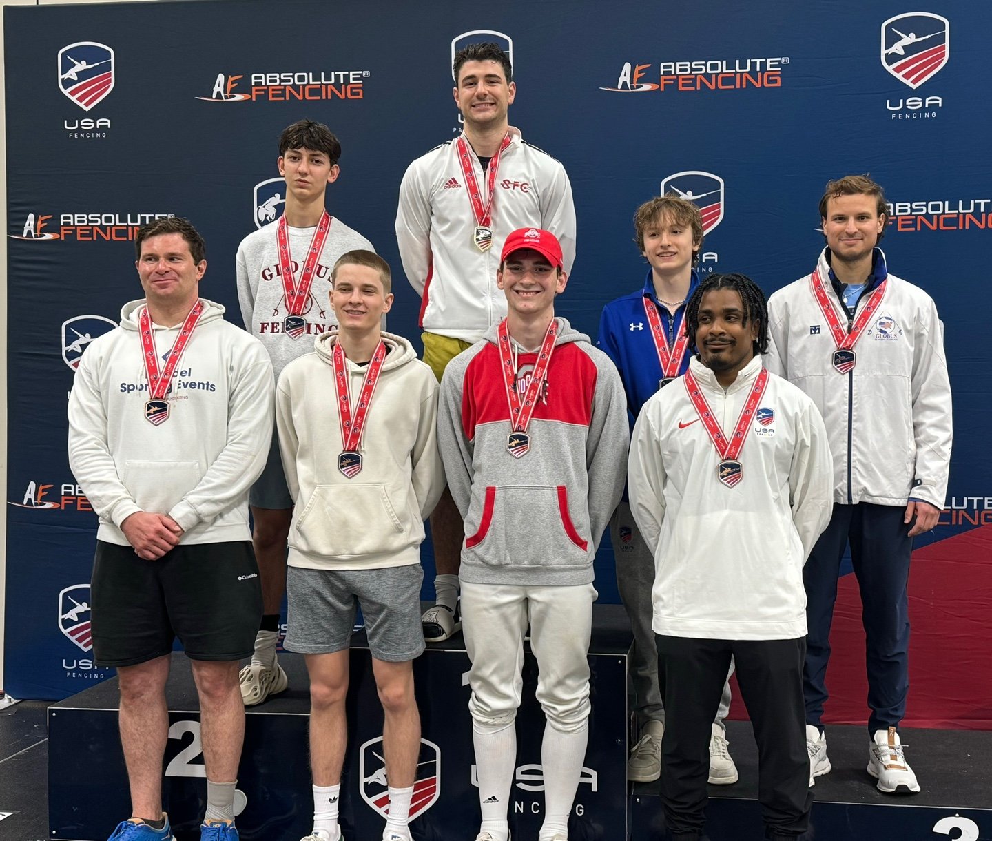 A group of ten male athletes on a winners' podium at a fencing competition, wearing medals and sportswear, with a backdrop displaying 'Absolute Fencing' and 'USA Fencing' logos.