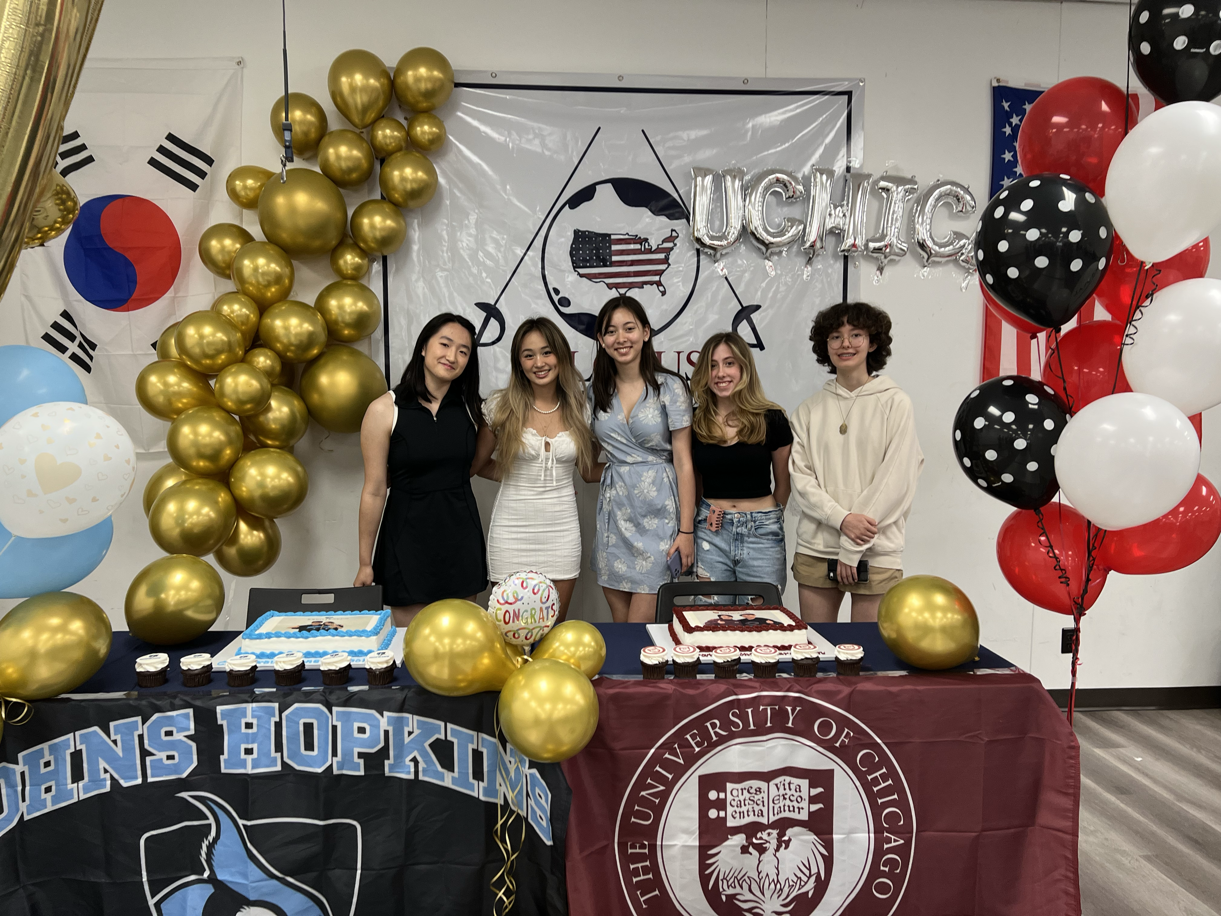 Five young women standing together at a celebration with balloons and cakes, in front of a backdrop with University of Chicago logos, South Korea and American flags, and golden, red, black, and white balloon decorations.