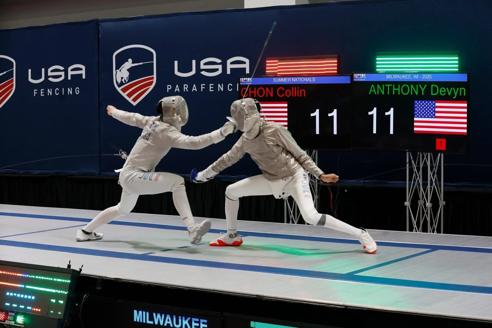 Two fencers in protective gear engaging in a match during the USA Summer Nationals in Milwaukee, Wisconsin. They are lunging at each other on a fencing piste with digital scoreboards behind them displaying their scores and names.