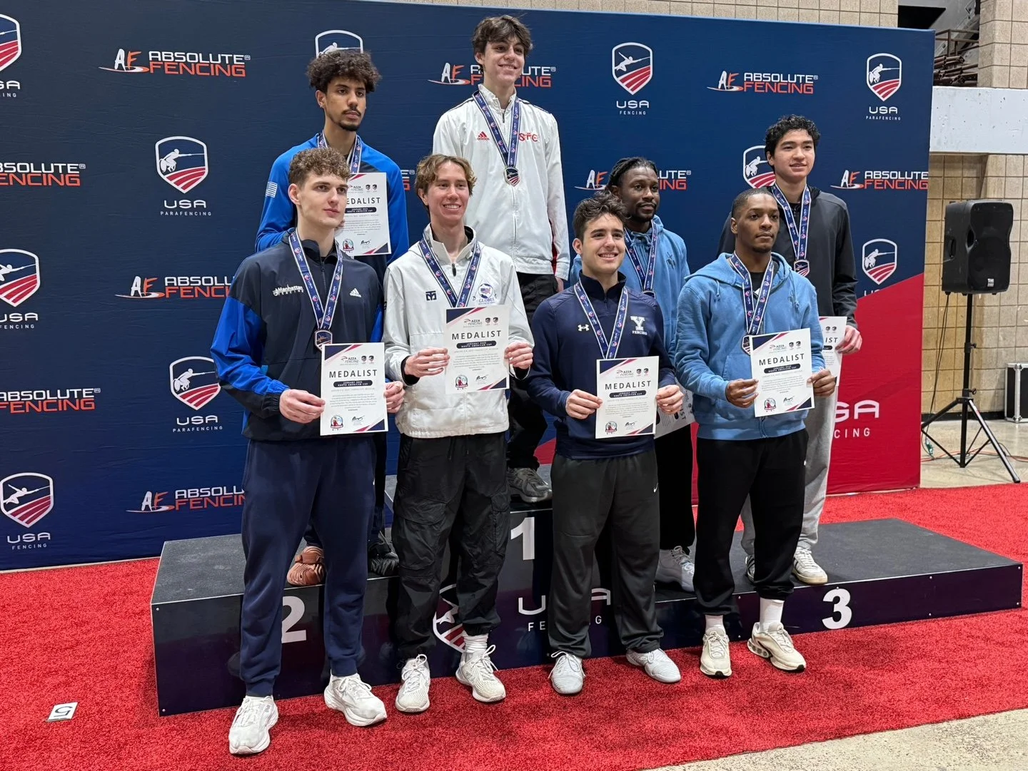 Group of eight young men standing on a winners' podium at a fencing competition, holding medals and certificates. The background features a blue and red backdrop with 'Absolute Fencing' and 'USA Parafencing' logos. The athletes are dressed in sportsw