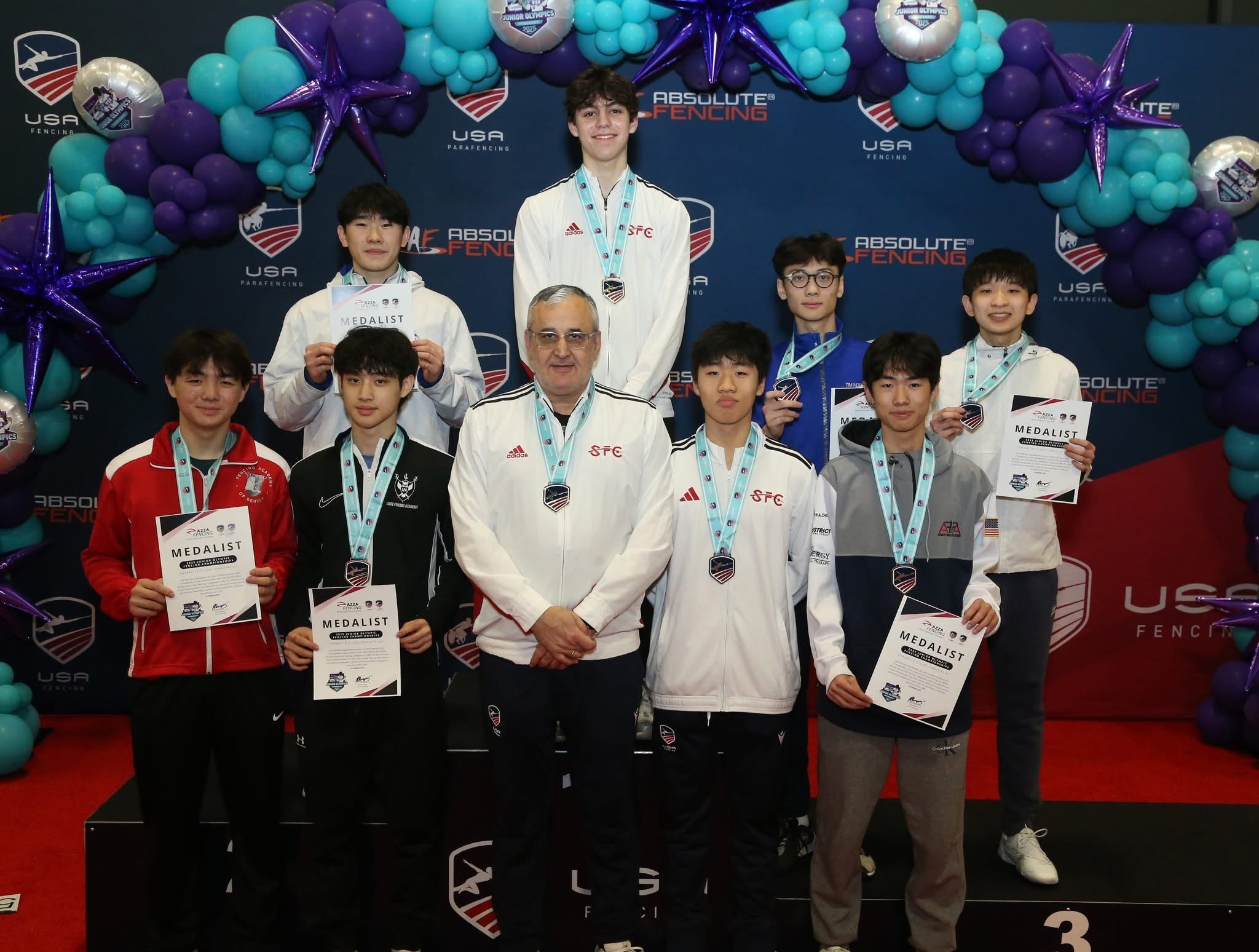 Group of young athletes and coach on podium at fencing awards ceremony, with medals and certificates, surrounded by balloon arch and backdrop with logos.