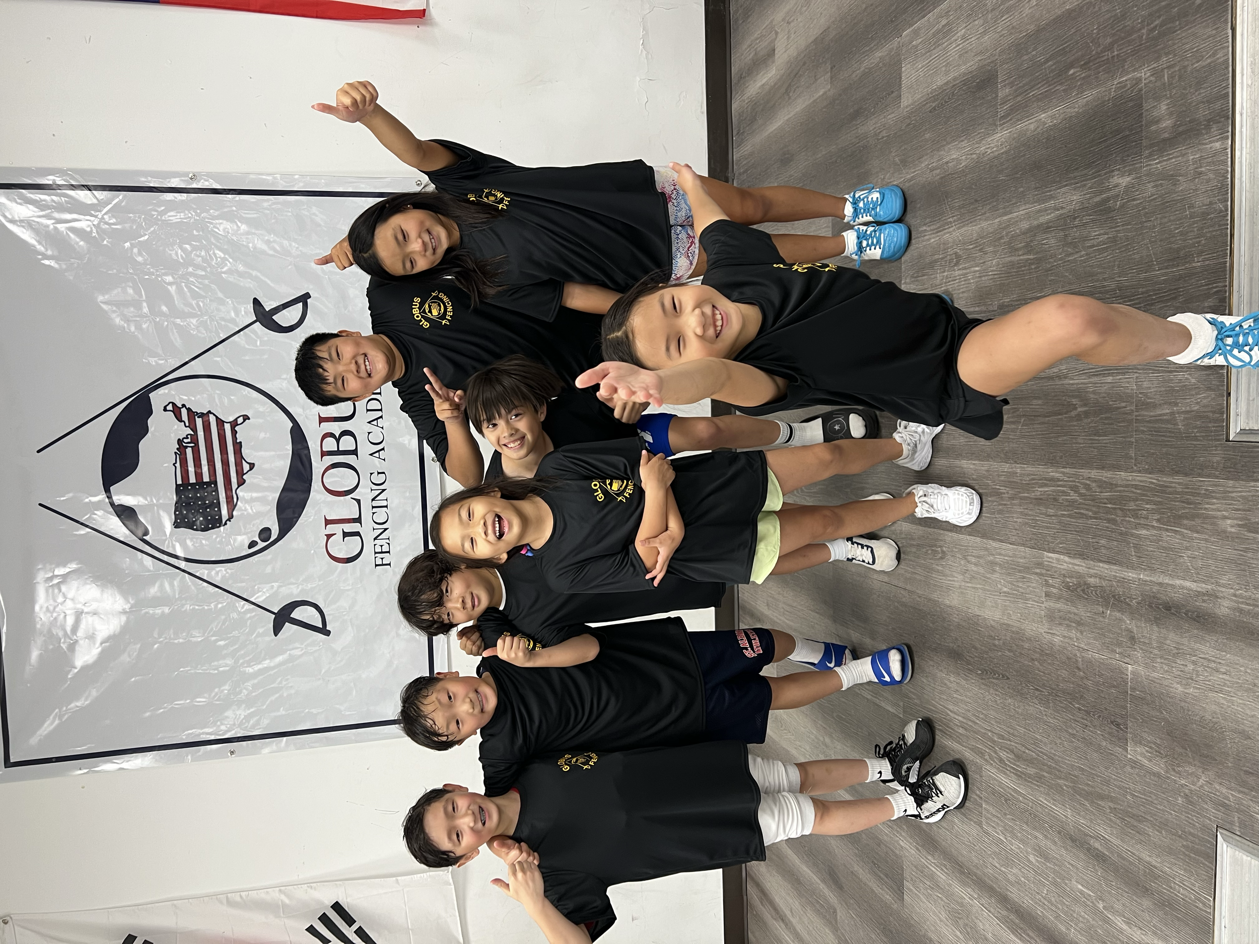 A group of children and their instructor at a fencing academy, wearing black team shirts, posing in front of a logo that reads 'GLOBUS FENCING ACADEMY' with a piggy bank and American flag design.
