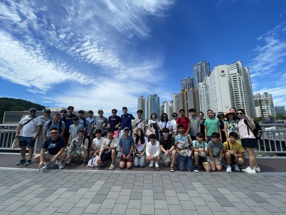 Group of people posing for a photo on a city bridge with tall buildings and a partly cloudy sky in the background.