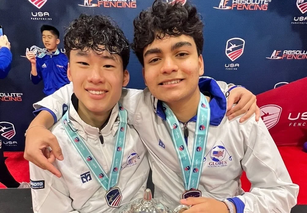 Two young male fencers with medals, smiling, with a third young male fencer in the background holding a medal, at a fencing competition