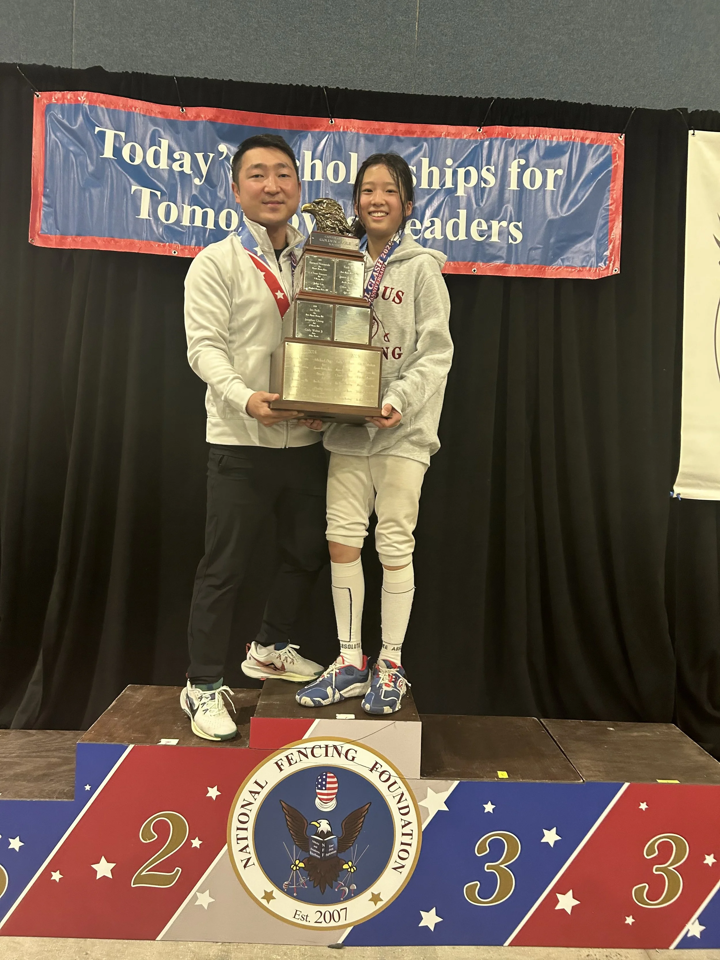 A young female athlete standing on the second-place podium, holding a large trophy, smiling with an adult male beside her at a fencing competition awards ceremony, with a banner in the background that reads 'Today's Scholarships for Tomorrow's Leader