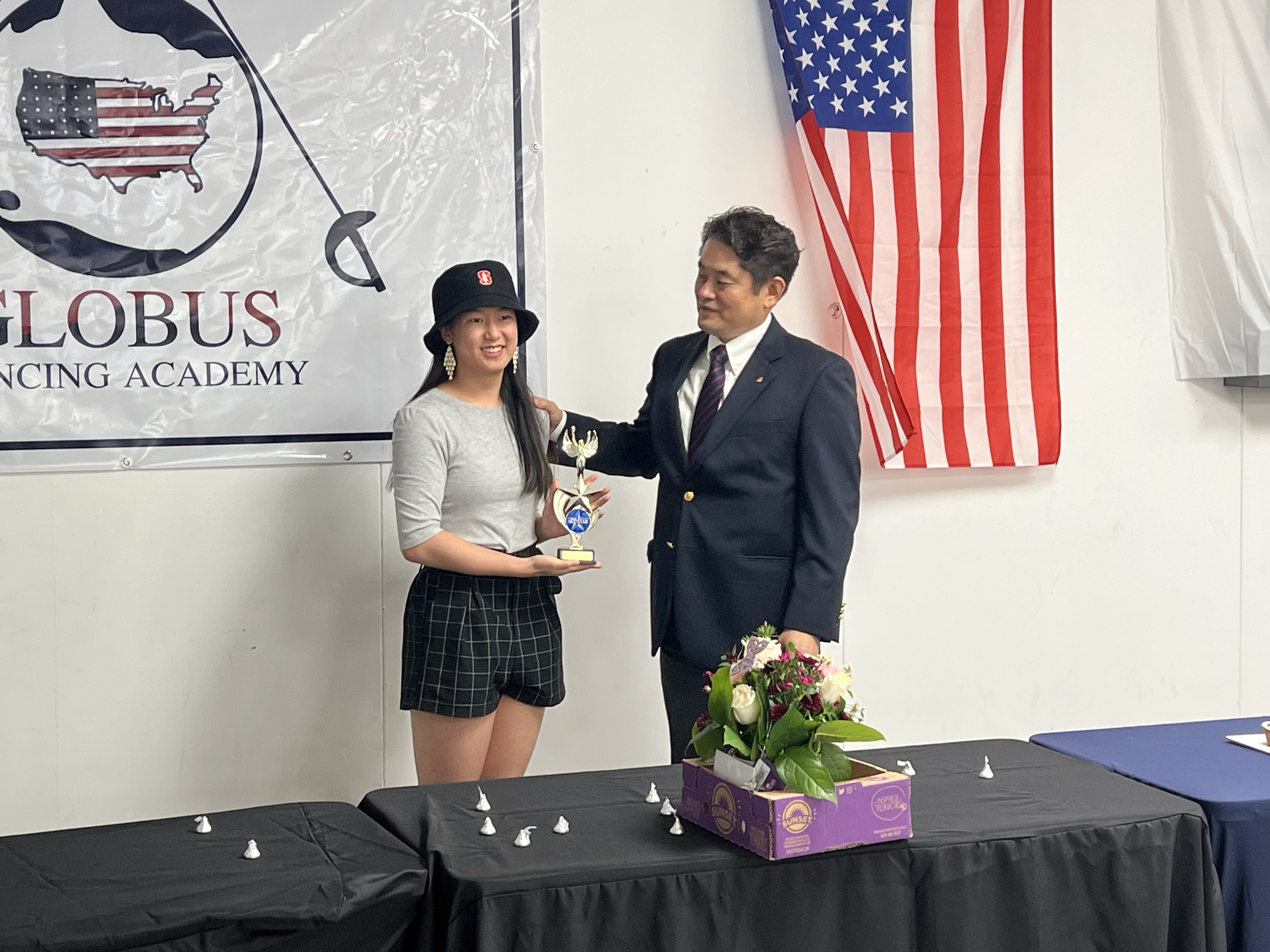 A young woman receiving a trophy from a man in a suit at a ceremony. The woman is smiling, wearing a black hat, gray shirt, and checkered shorts. The man is in a dark suit with a tie. There is a large American flag on the wall and a banner with a map