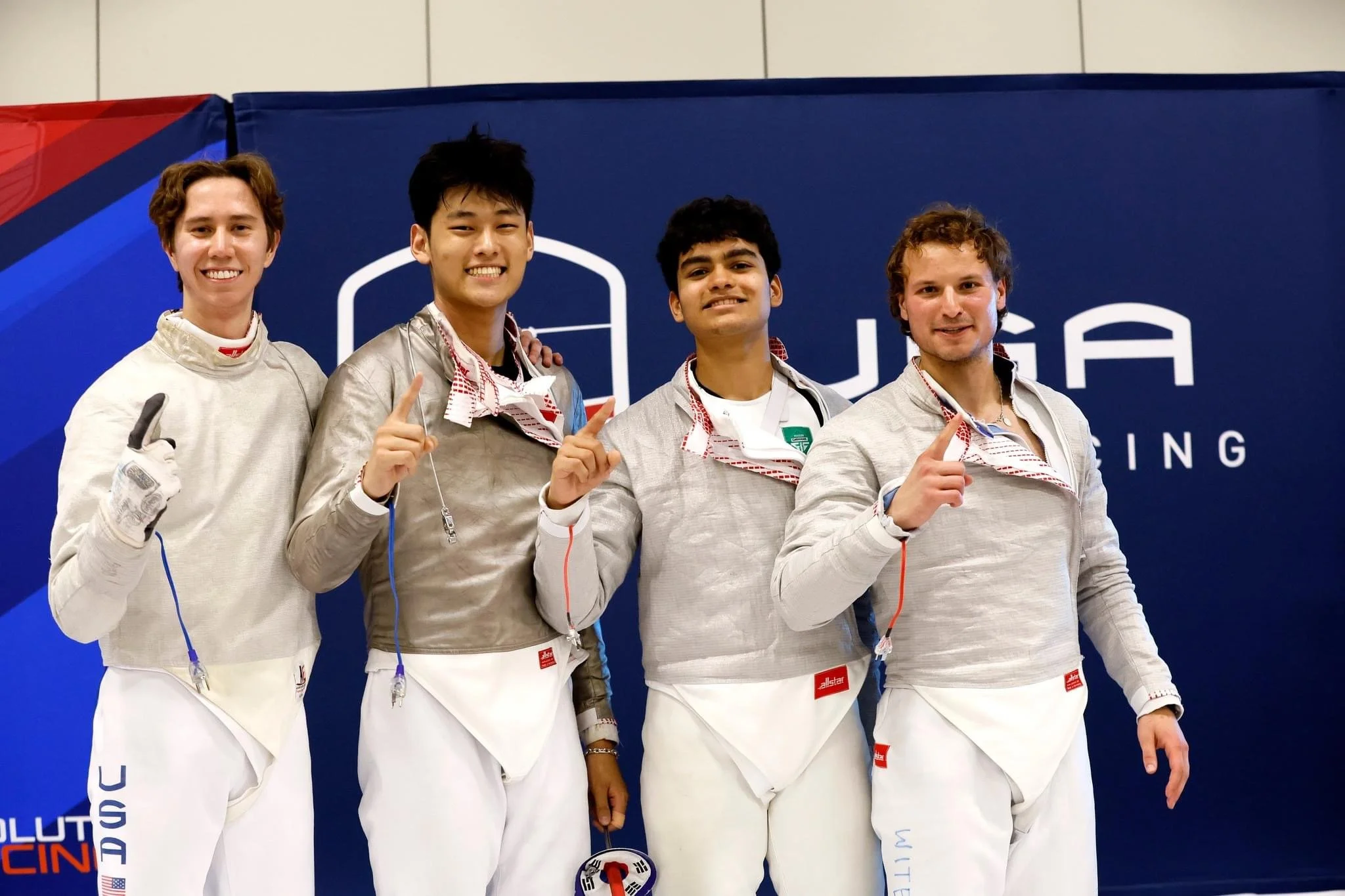 Four male fencers in white and gray uniforms and fencing gear stand together, smiling, in front of a blue backdrop with fencing logo and text.