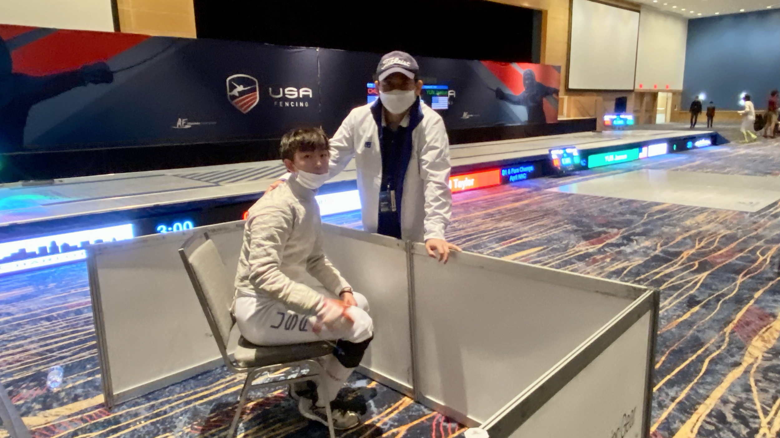 Two people, a young man sitting and an older man standing, wear masks at a fencing event in an indoor arena, with a USA fencing sign and electronic scoreboard in the background.