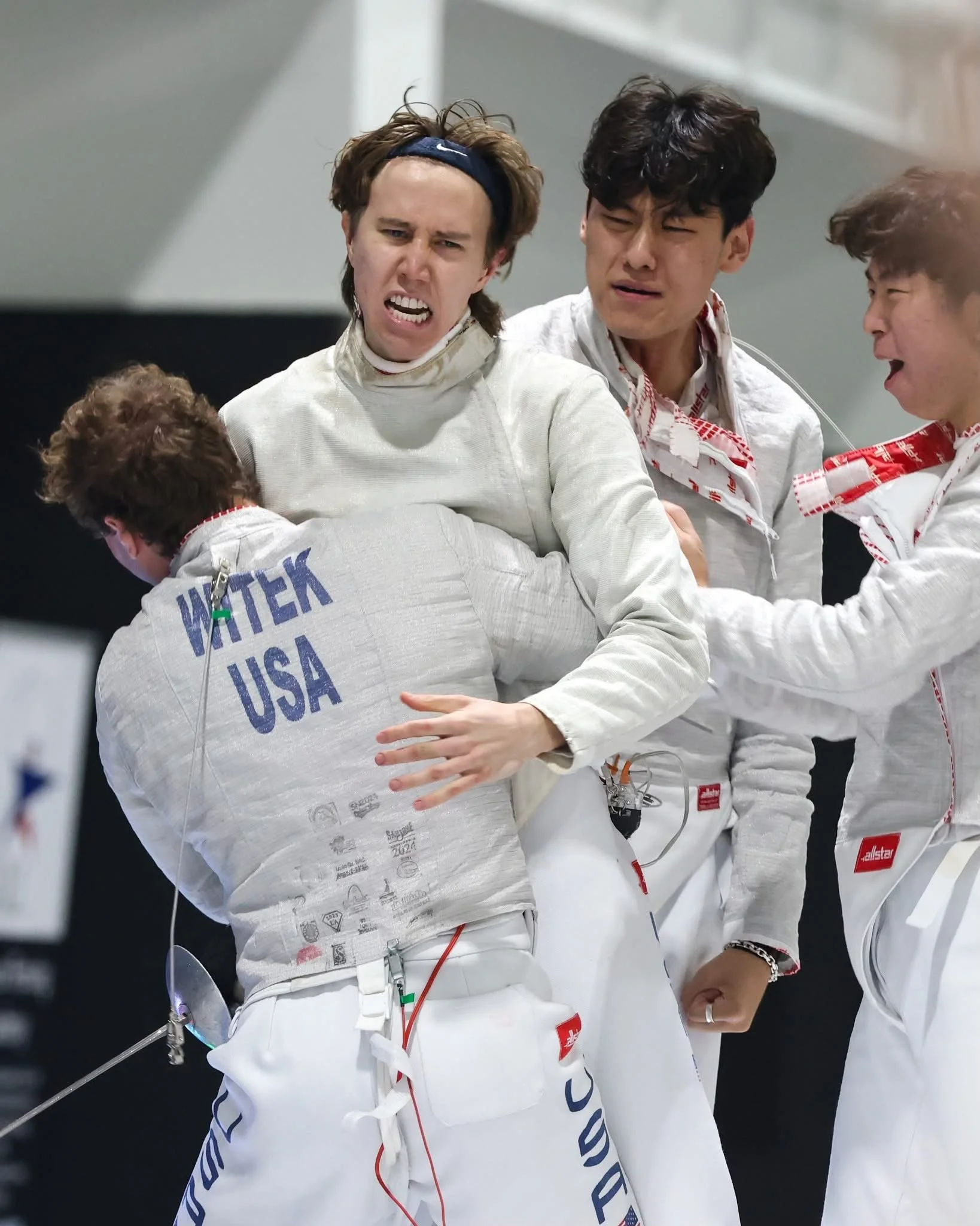 Celebrating athletes in fencing gear, embracing after a match, with 'WATER USA' on the back of one athlete's jacket, at a competition.