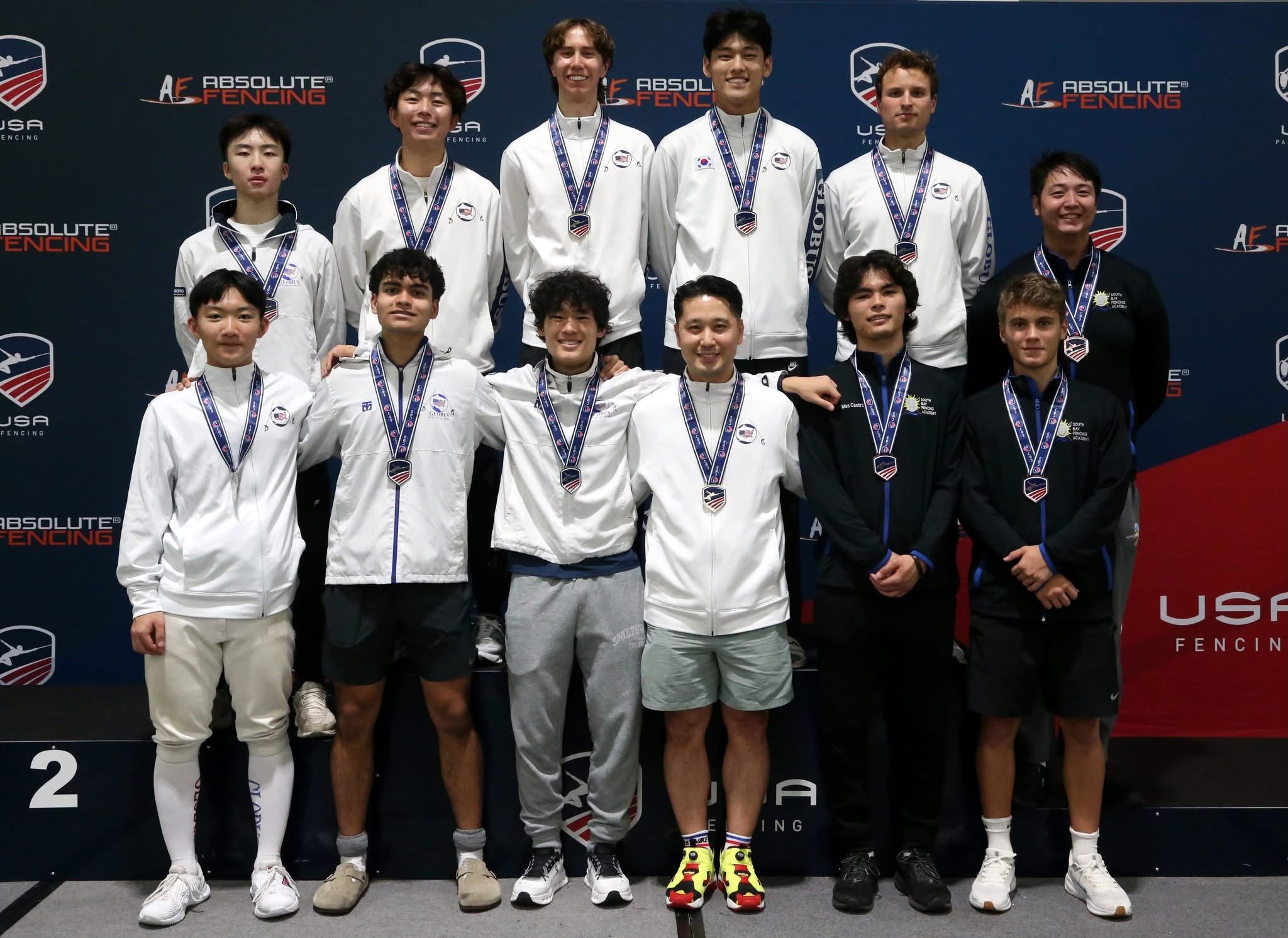 A group of eleven young athletes standing on a winners' podium at a fencing competition, all wearing medals and sports uniforms, with a backdrop displaying the 'Absolute Fencing' and 'USA Fencing' logos.