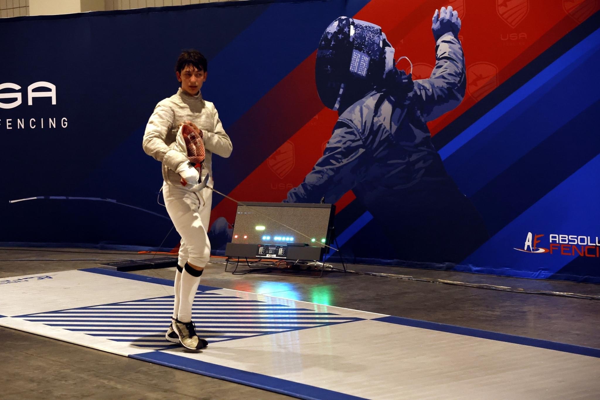 A young male fencer dressed in a white fencing uniform holding a fencing sword, standing on a fencing strip with a digital score display and electronic scoring system behind him, in an indoor fencing arena with a large mural of a fencer in the backgr