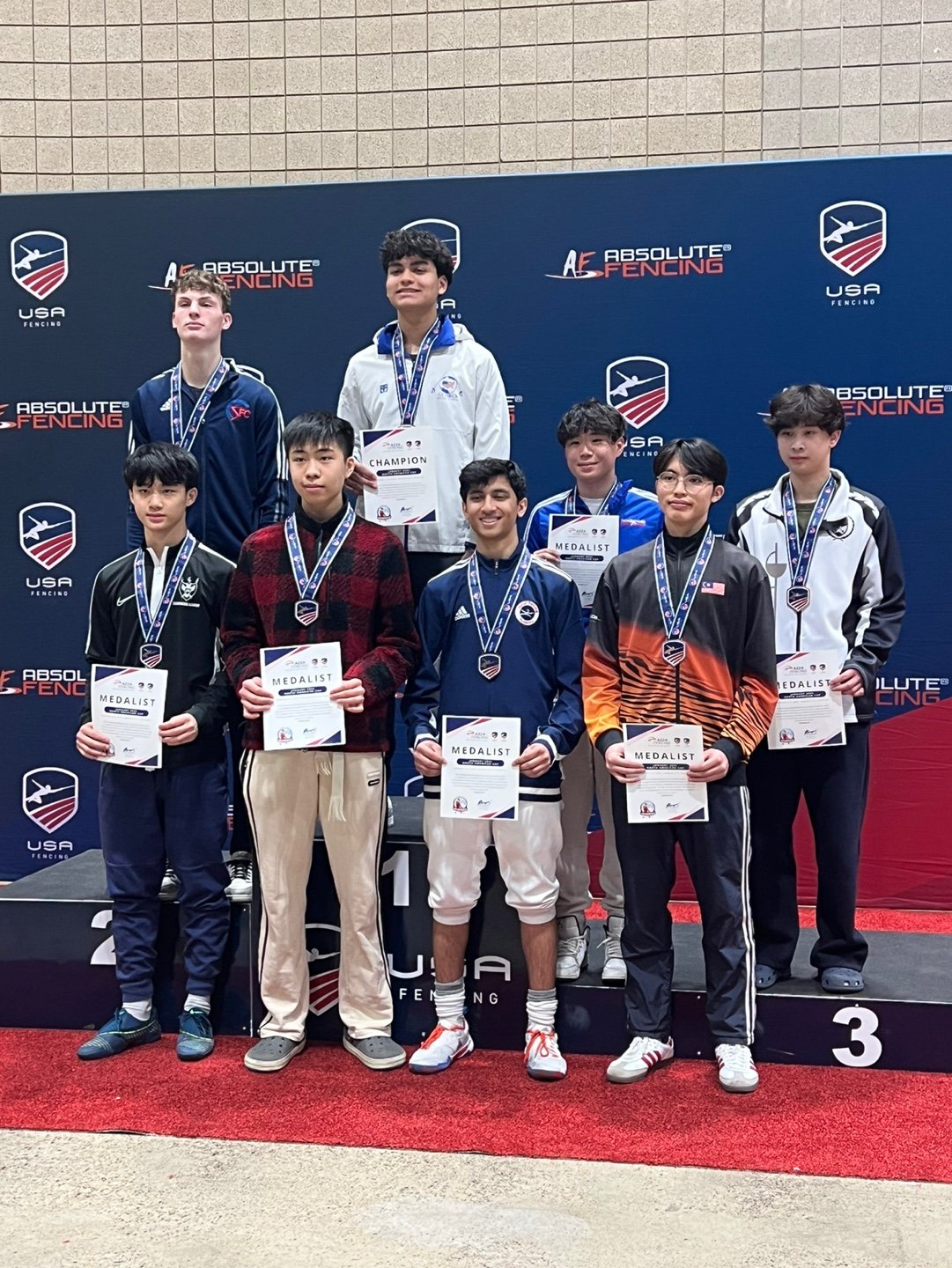 Young male fencers on a winners' podium holding certificates and medals, with a blue backdrop displaying the USA Fencing logo and 'Absolute Fencing' branding.