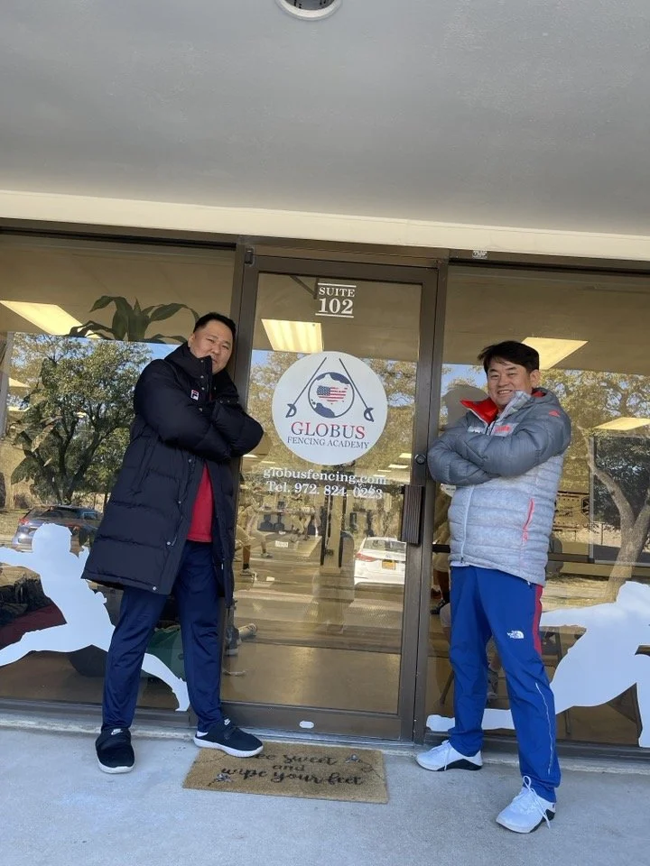 Two men standing outside the entrance of Globus Fencing Academy, a fencing school, in front of the glass door with the academy's sign and logo, crossing their arms and posing for the photo.